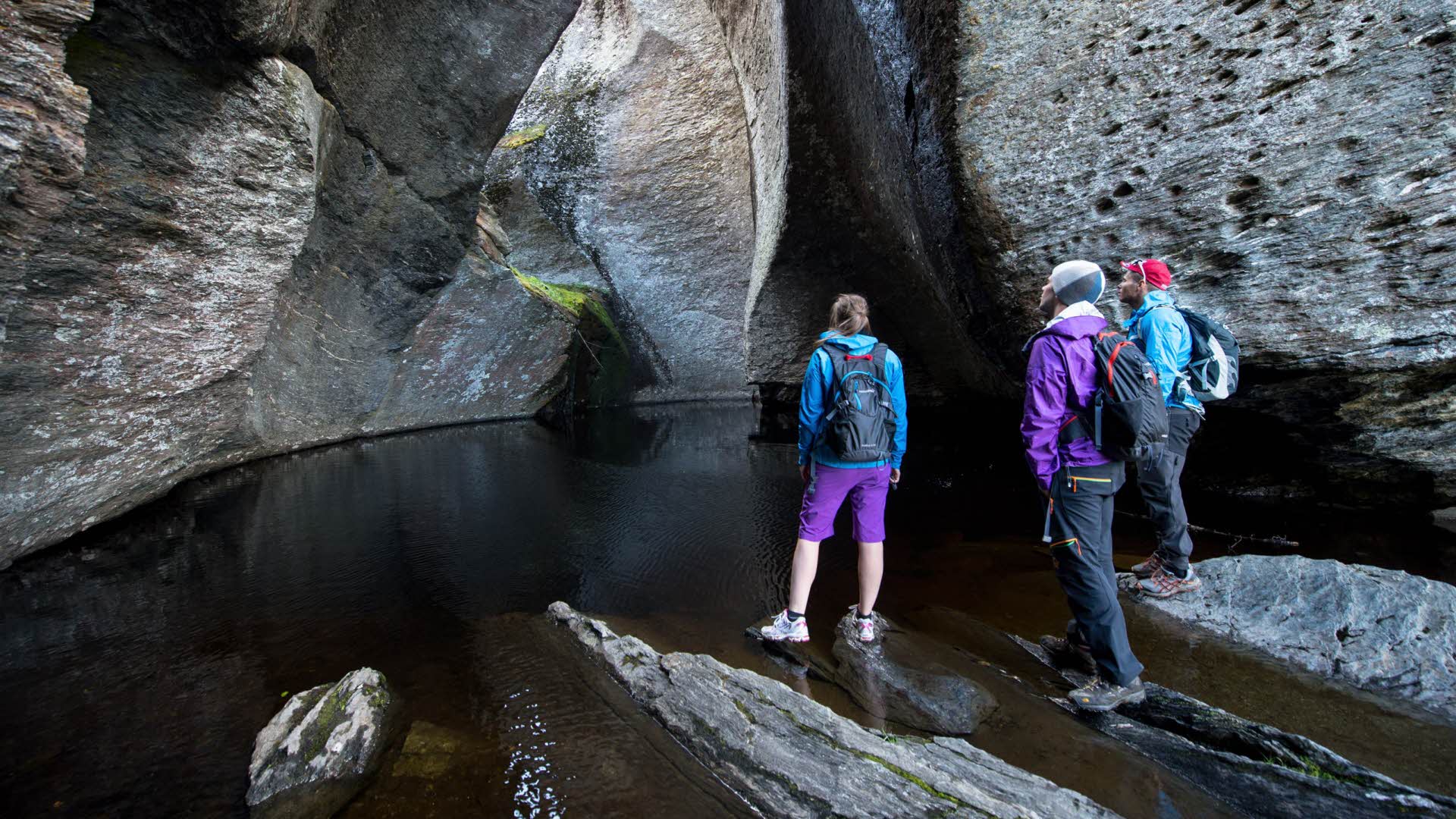 3 turgåere i står ved foten av det sorte vannet av Vetlehelvete i Aurlandsdalen og ser på de fasinerende steinformasjonene