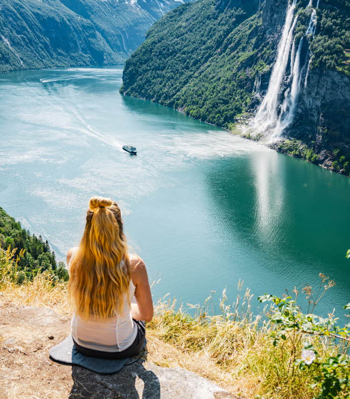 A woman sitting on a plateau above the Geirangerfjord. A boat sailing and a cascading waterfall below her.