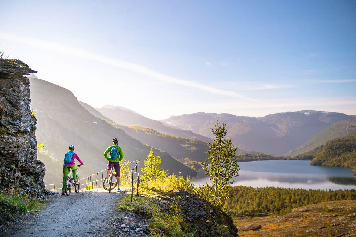 To personer står over syklene sine ved en stor stein og ser utover et vann og fjell på Rallarvegen.
