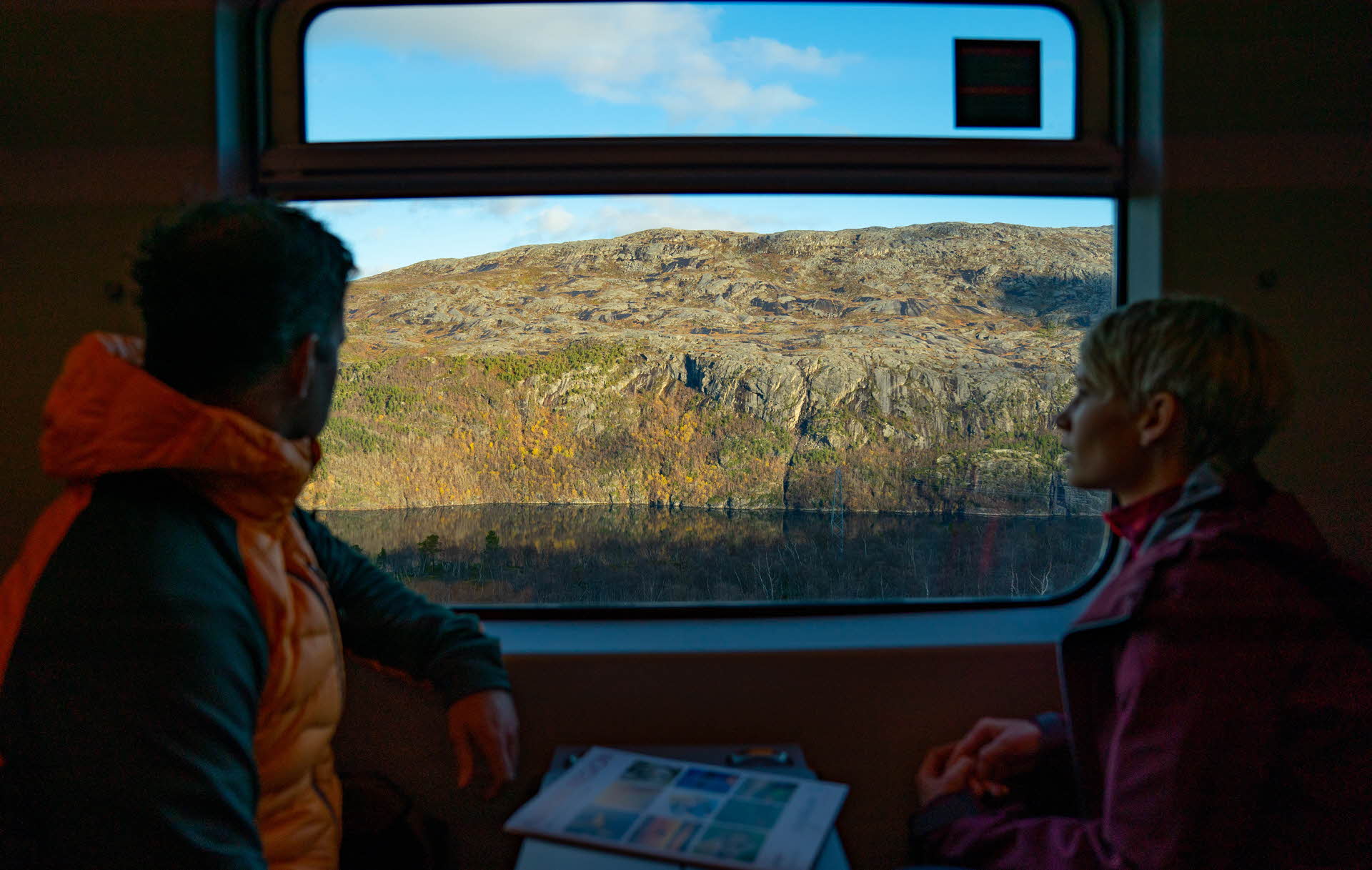 An mann og en kvinne sitter mot hverandre på et tog og ser ut av vinduet mot fjell og fjord.