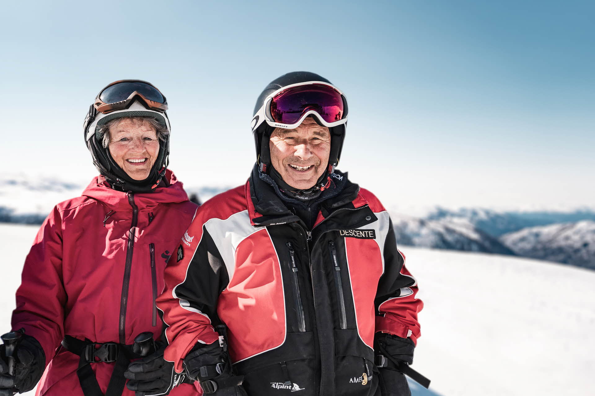 Una pareja de personas mayores con chaquetas rojas y negras y cascos de pie en las pistas de Myrkdalen sonriendo a la cámara.