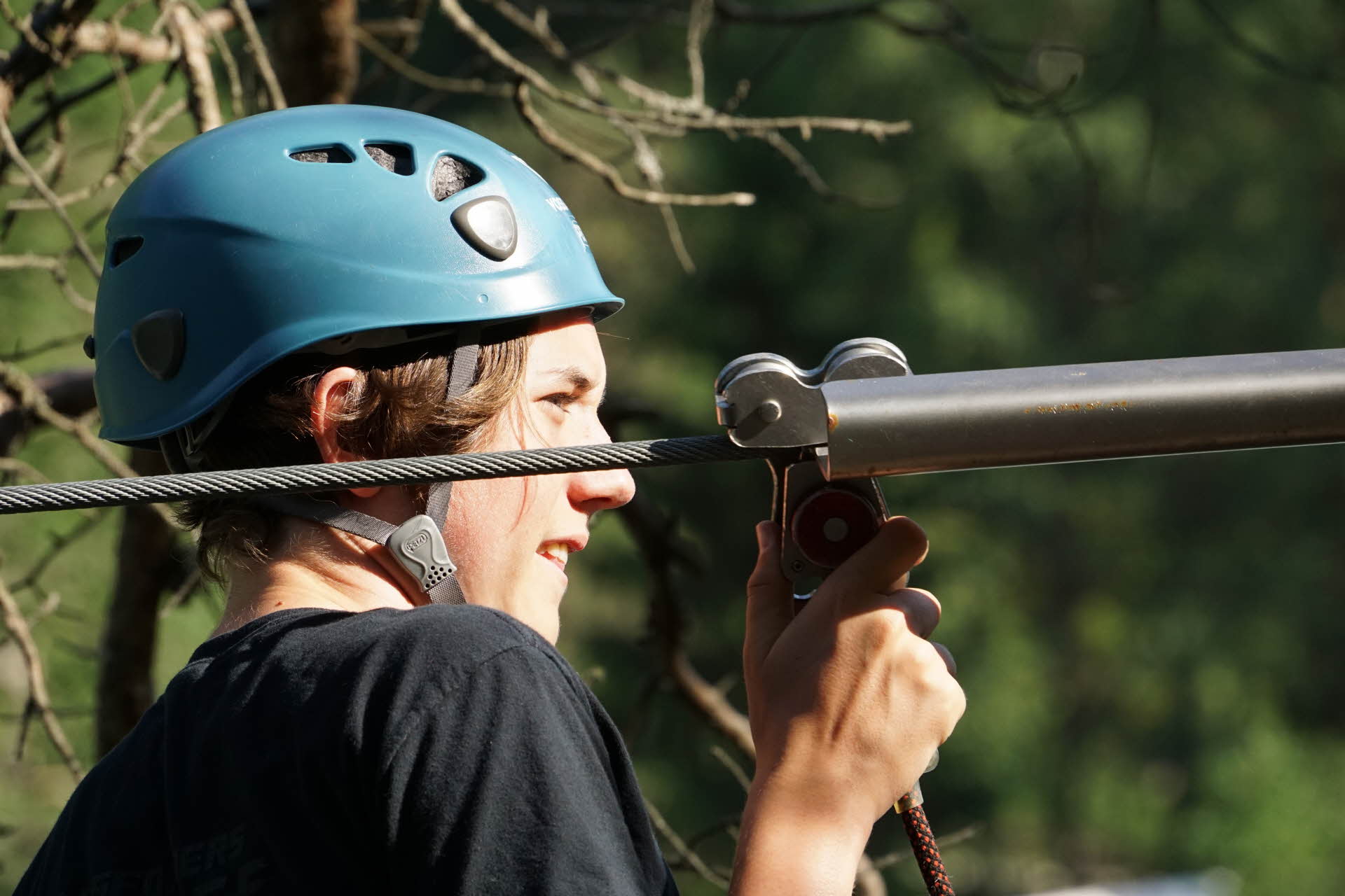En ungdom i blå hjelm fester sikkerhetskarabinen på vaier i Voss klatrepark.