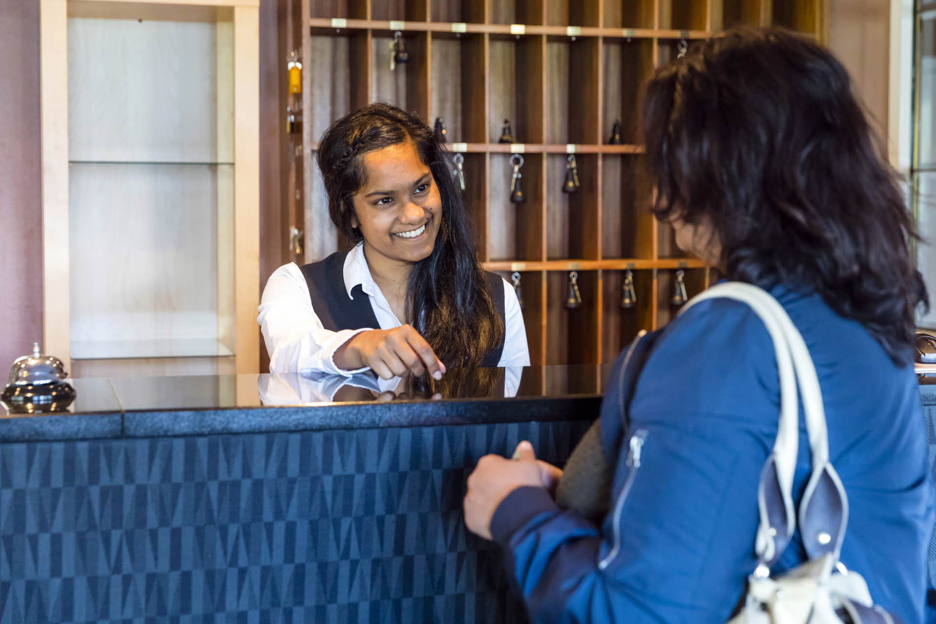 Una mujer con chaqueta azul en una recepción. Una recepcionista sonriente entregando una llave.