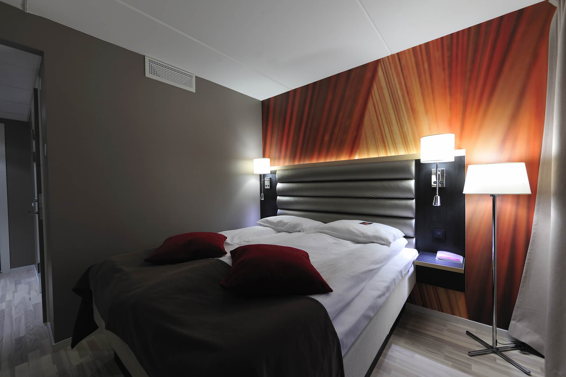 Doppelbett, Lampen, eine bunte Wand und rote Zierkissen. 