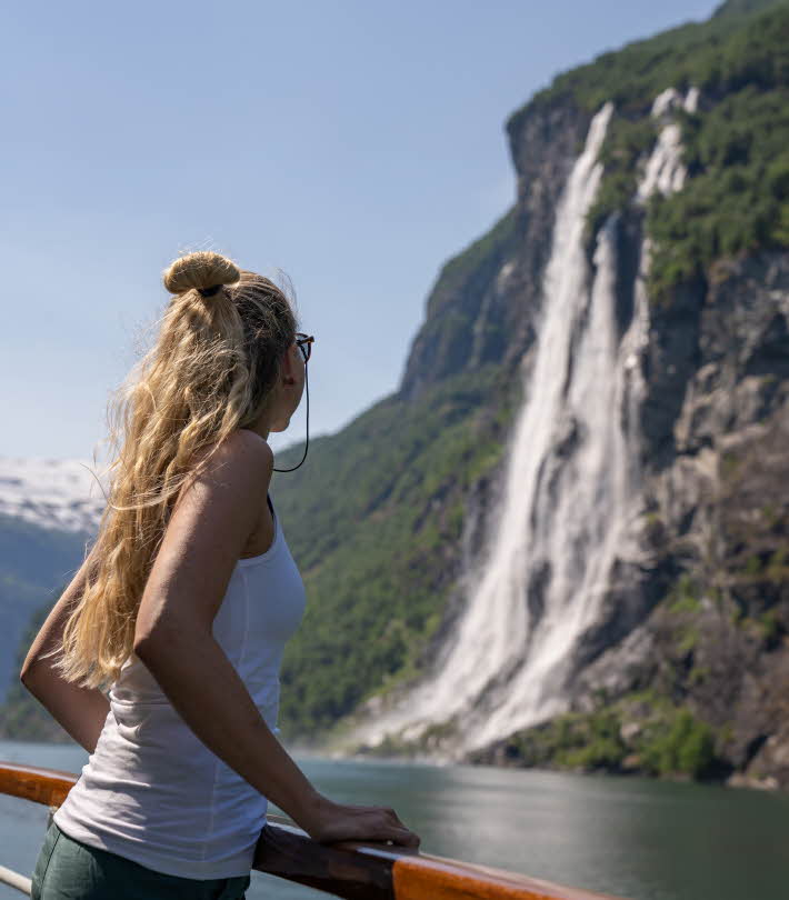 A woman with long blond hair looks at the Seven Sisters Waterfall on the Geirangerfjord