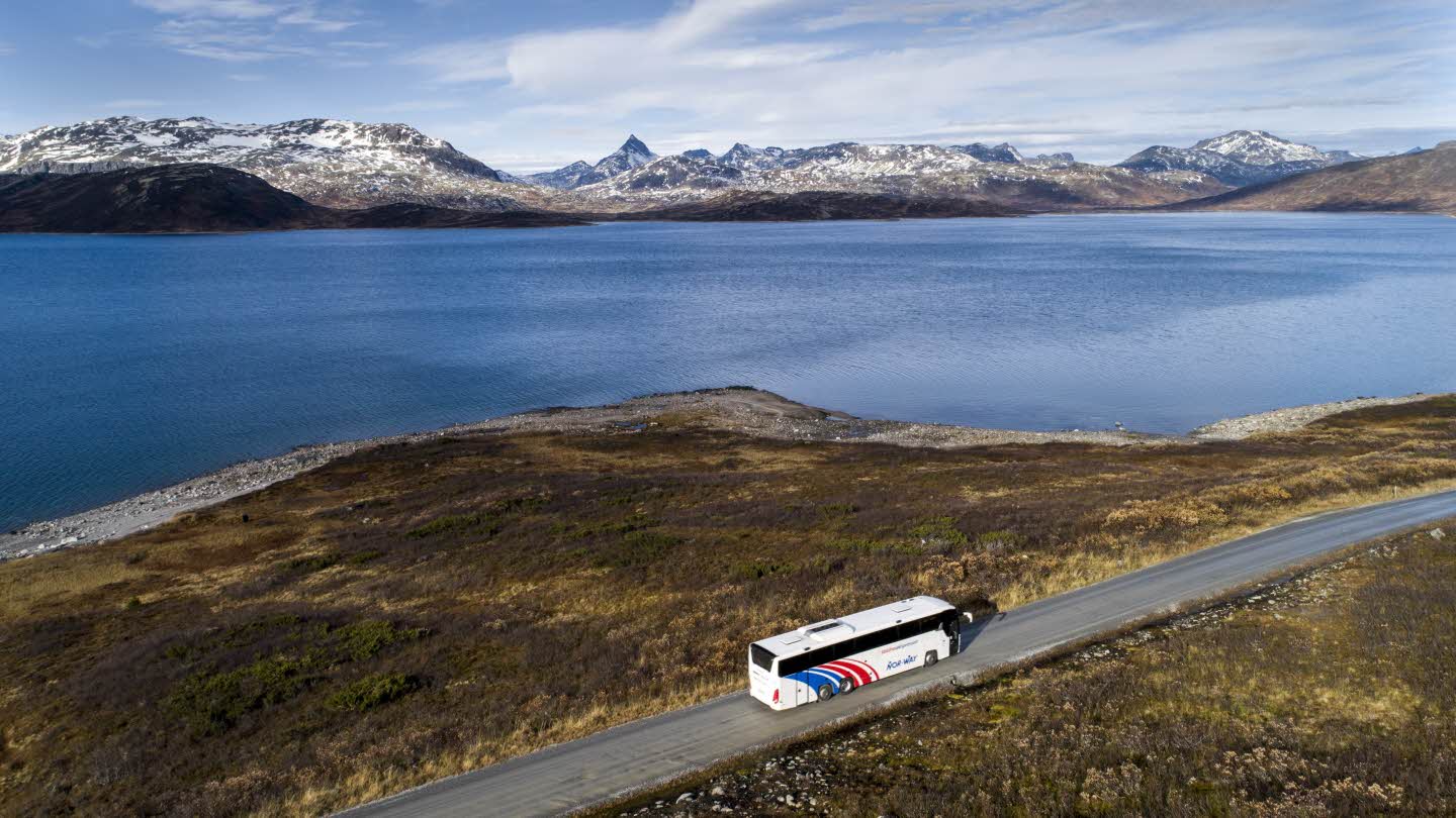 A bus driving on the gravel road to Eidsbugarden. Peaks of Jjotunheimen National Park seen behind a lake. 
