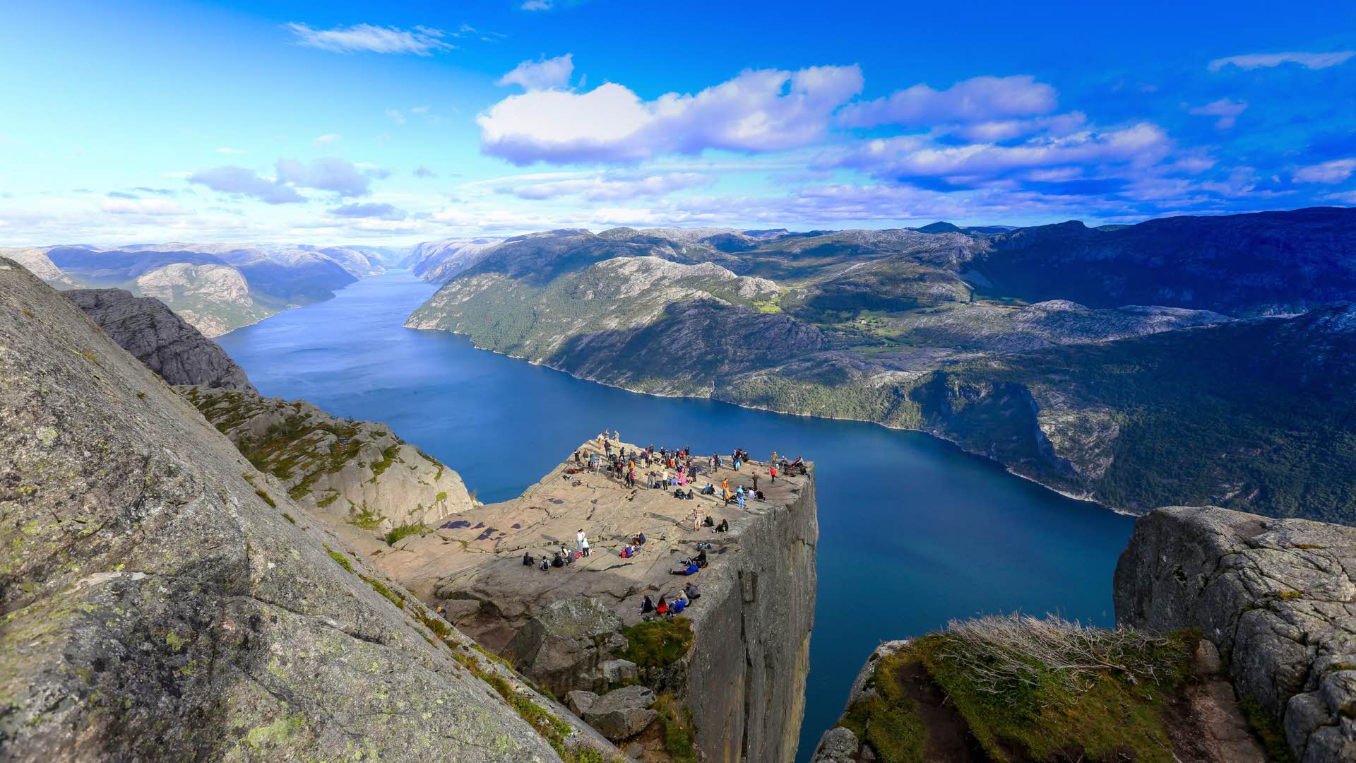 Panorama view from above Pulpit Rock, crowd of people standing near edge. Lysefjord can be seen as stretching out to infinity