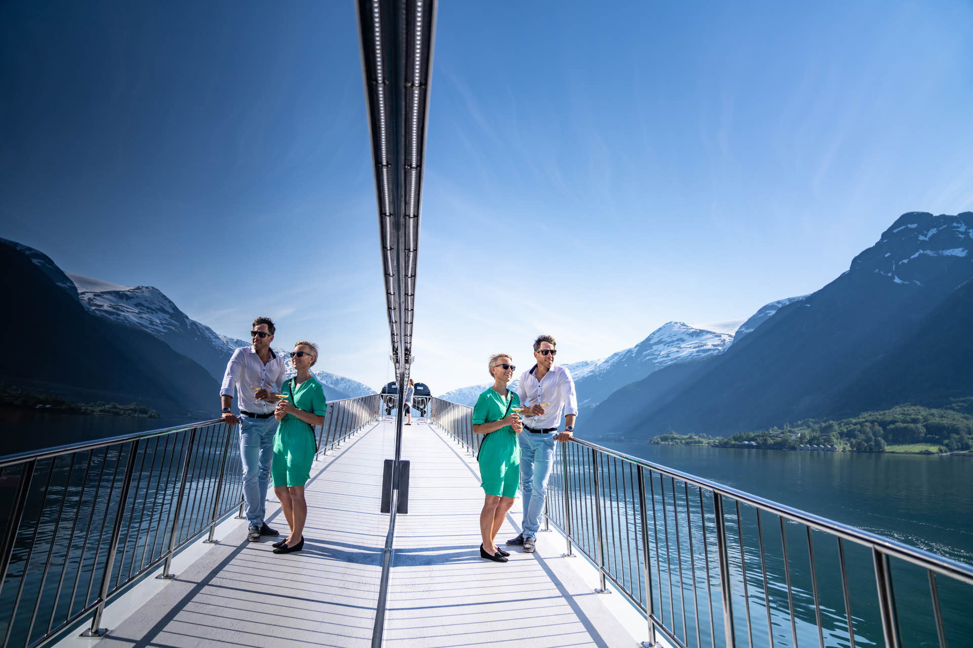 Un homme et une femme en vêtements d’été debout sur le pont d’un bateau dans le Hardangerfjord. Les fenêtres reflètent le paysage.