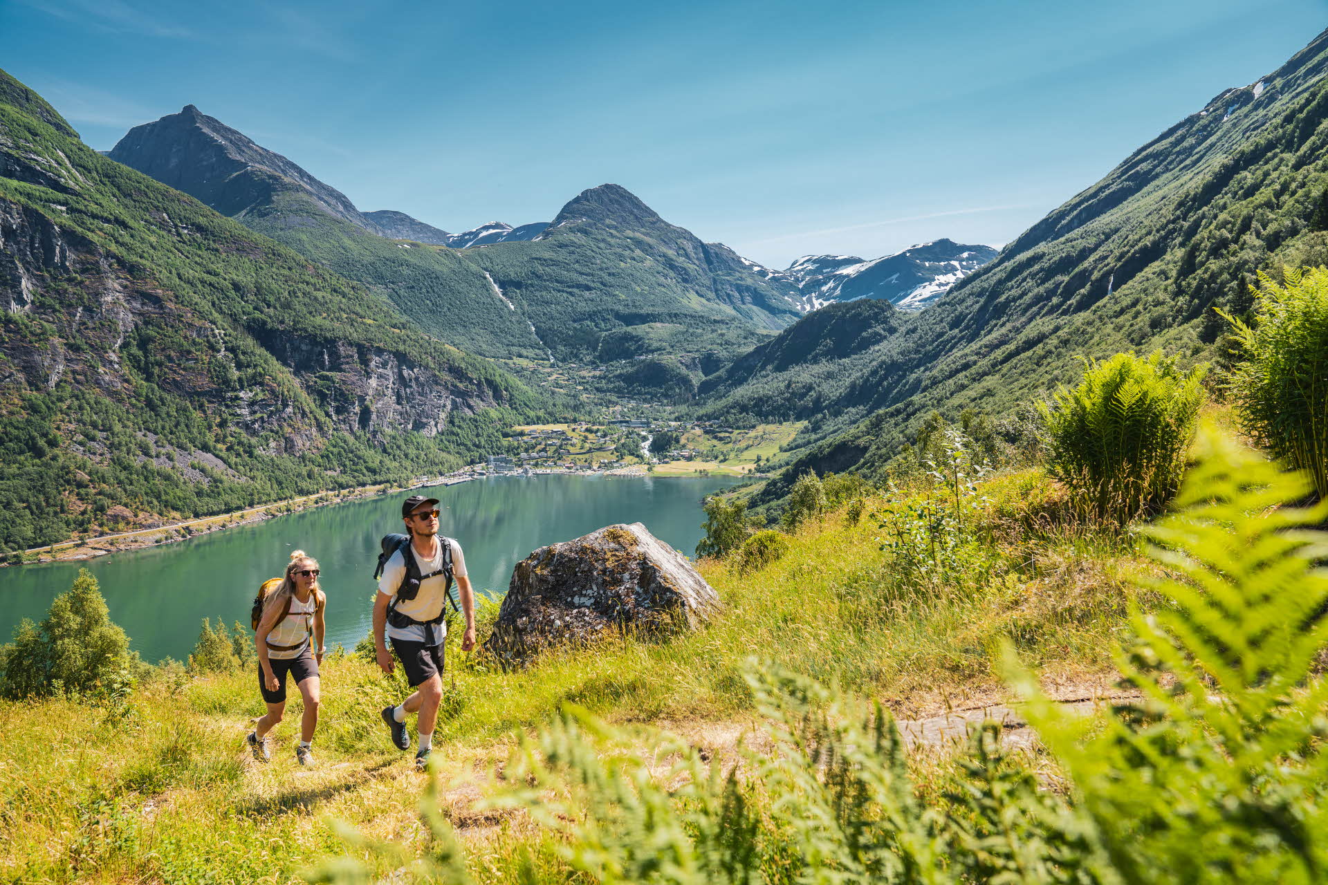 To personer går oppover en grønn bakke over Geirangerfjorden. Geiranger og fjell rundt.