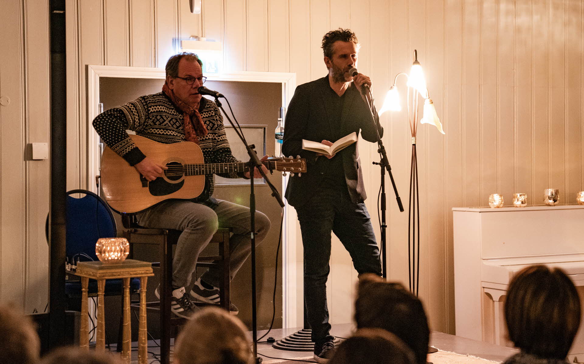 A man reading from a book into a microphone and another singing and playing guitar on a stage at Fretheim Hotel.