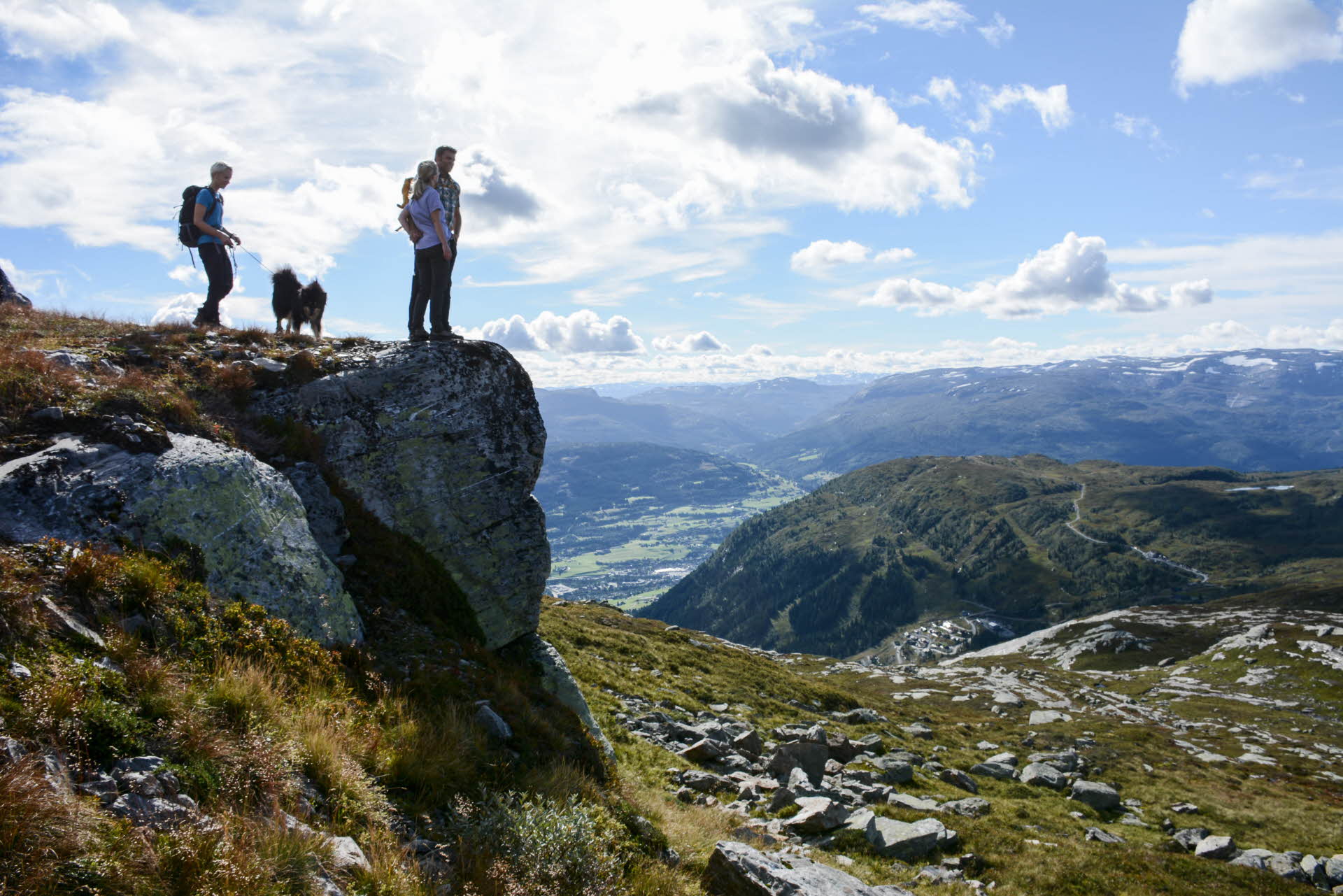 Une femme avec un chien et un couple en tenue de randonnée admirent la vue imprenable depuis la montagne Hangurstoppen au-dessus de Voss en été