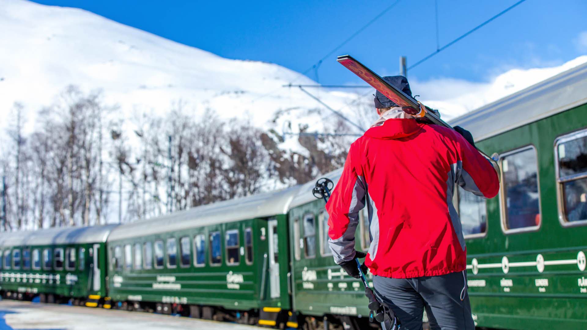 Un homme portant des skis sur ses épaules à côté d’un train du chemin de fer de Flåm, sur fond de montagnes enneigées.