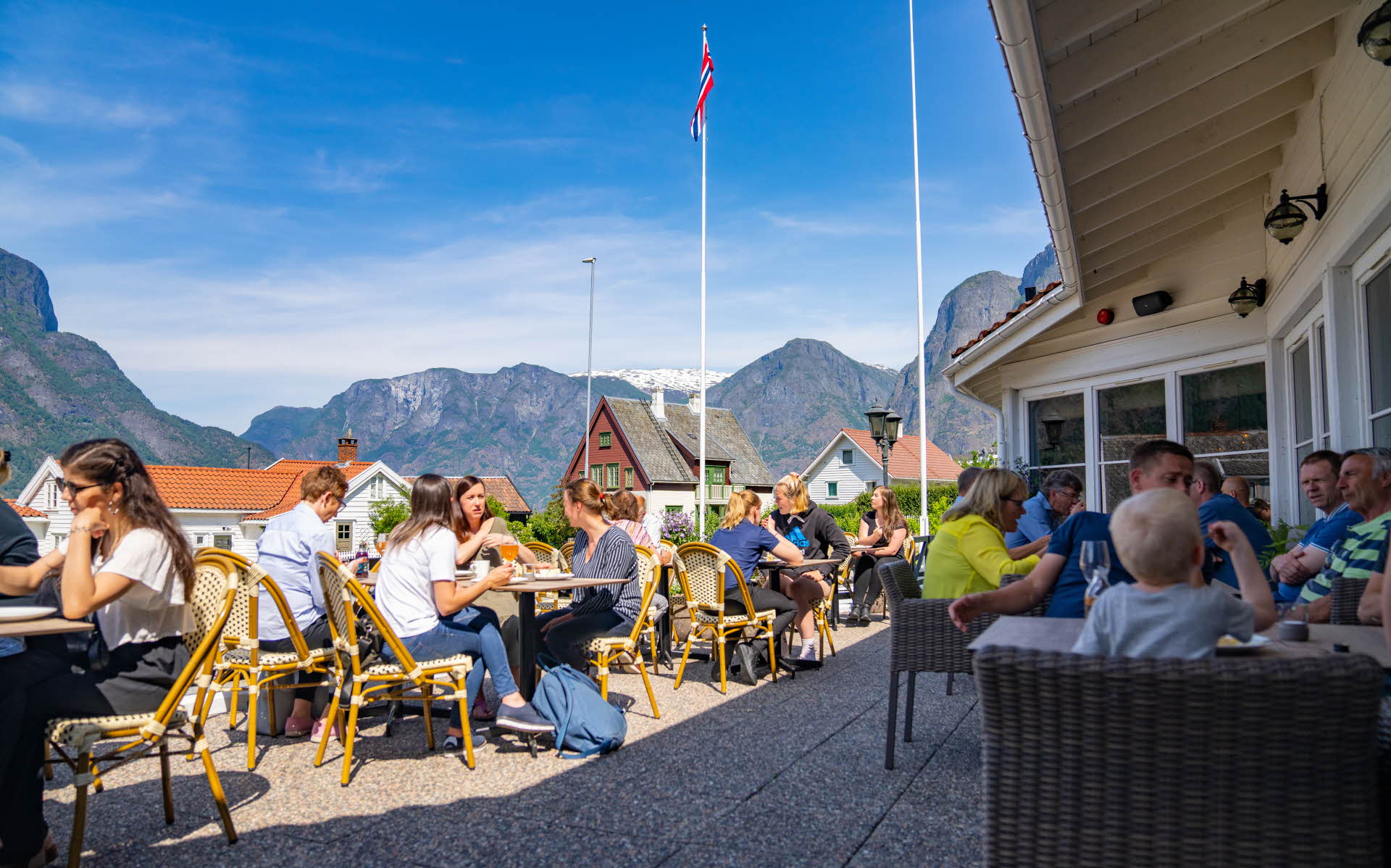 People sitting around tables on the terrace of the Hotel Aurlandsfjord. Houses and tall mountains in the background.