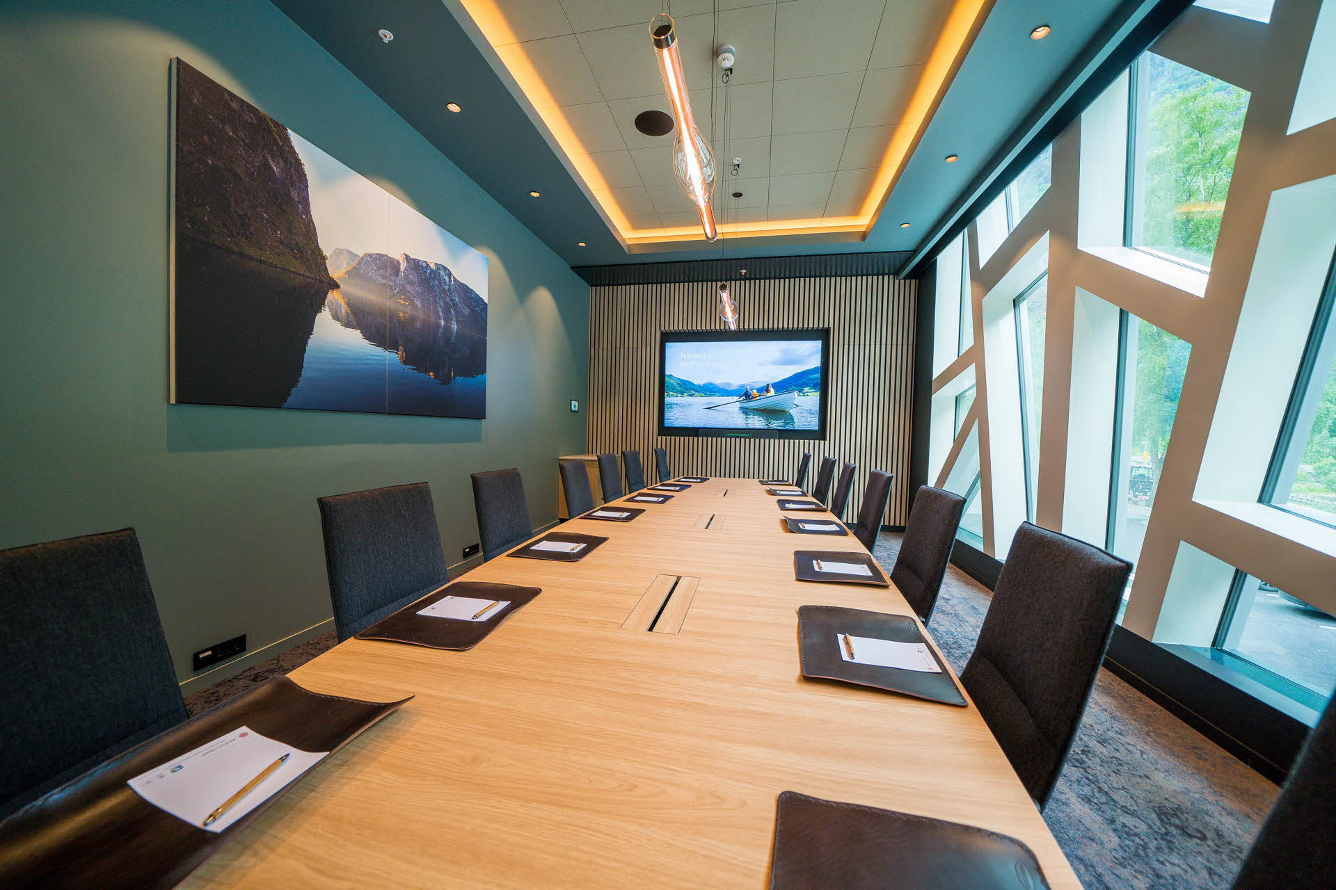 A meeting room with a long table and leather chairs. Large windows, picture and a screen on the walls. 