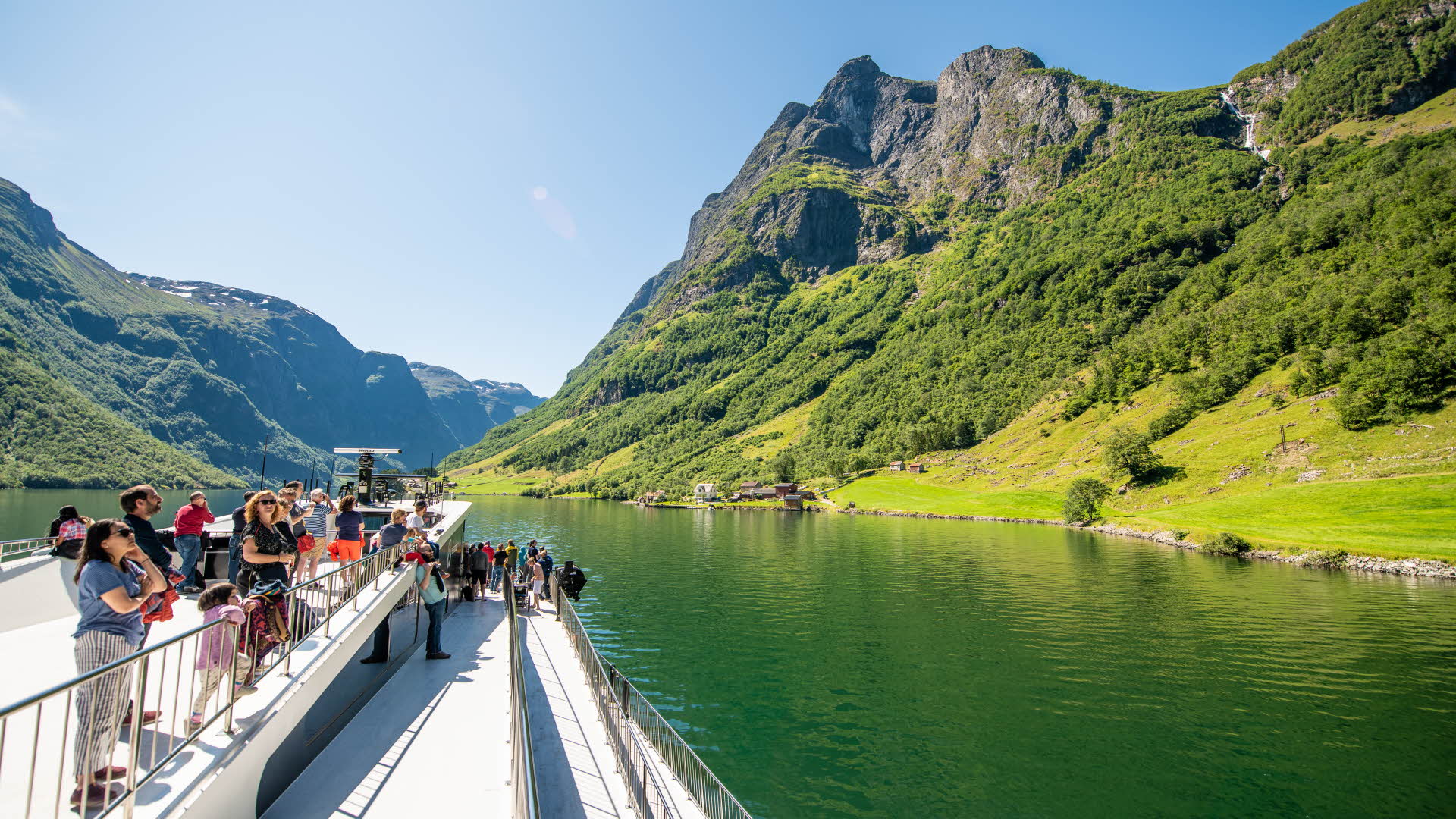 People standing on a roof deck cruising on the Nærøyfjord surrounded by steep, green mountains.