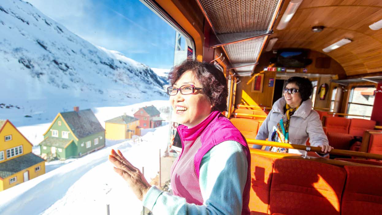 Zwei lächelnde asiatische Damen stehen in der Flåmbahn und blicken auf den Schnee und die bunten Häuser in Myrdal