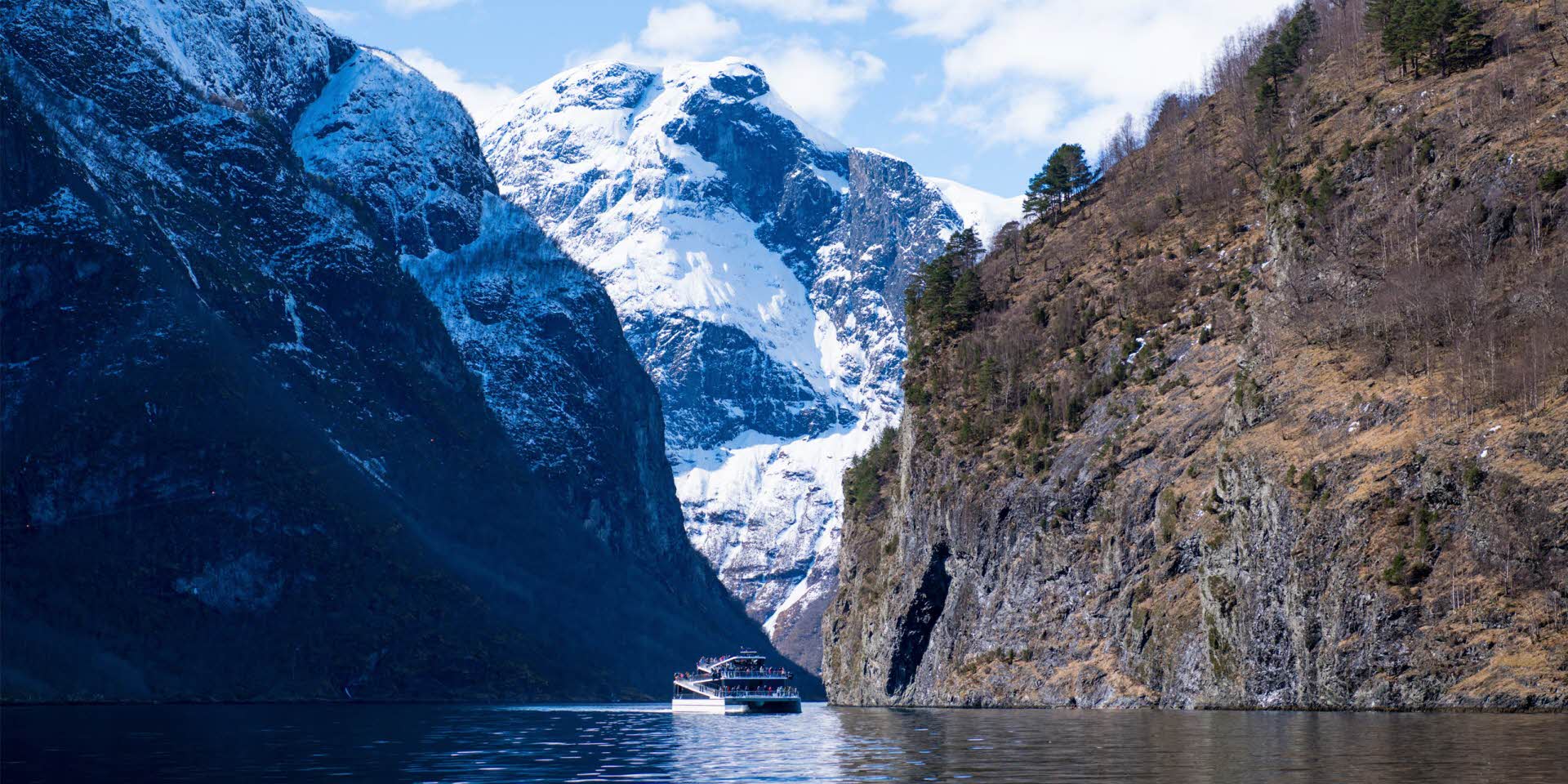 Le bateau Future of the Fjords naviguant dans le Nærøyfjord en fin d’hiver avec des montagnes enneigées et des personnes debout sur le pont