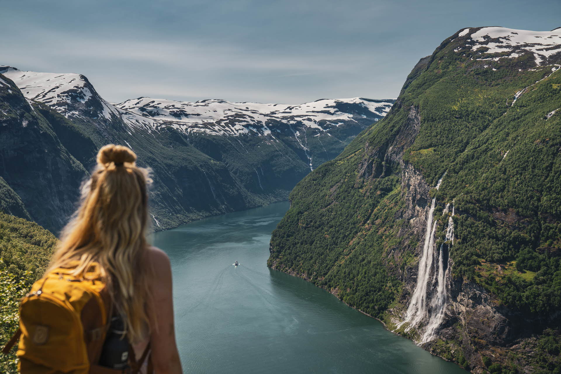 Eine Frau mit langem blondem Haar und einem gelben Rucksack blickt hinunter auf den Geirangerfjord, schneebedeckte Berge und der Wasserfall Die Sieben Schwestern im Hintergrund. 