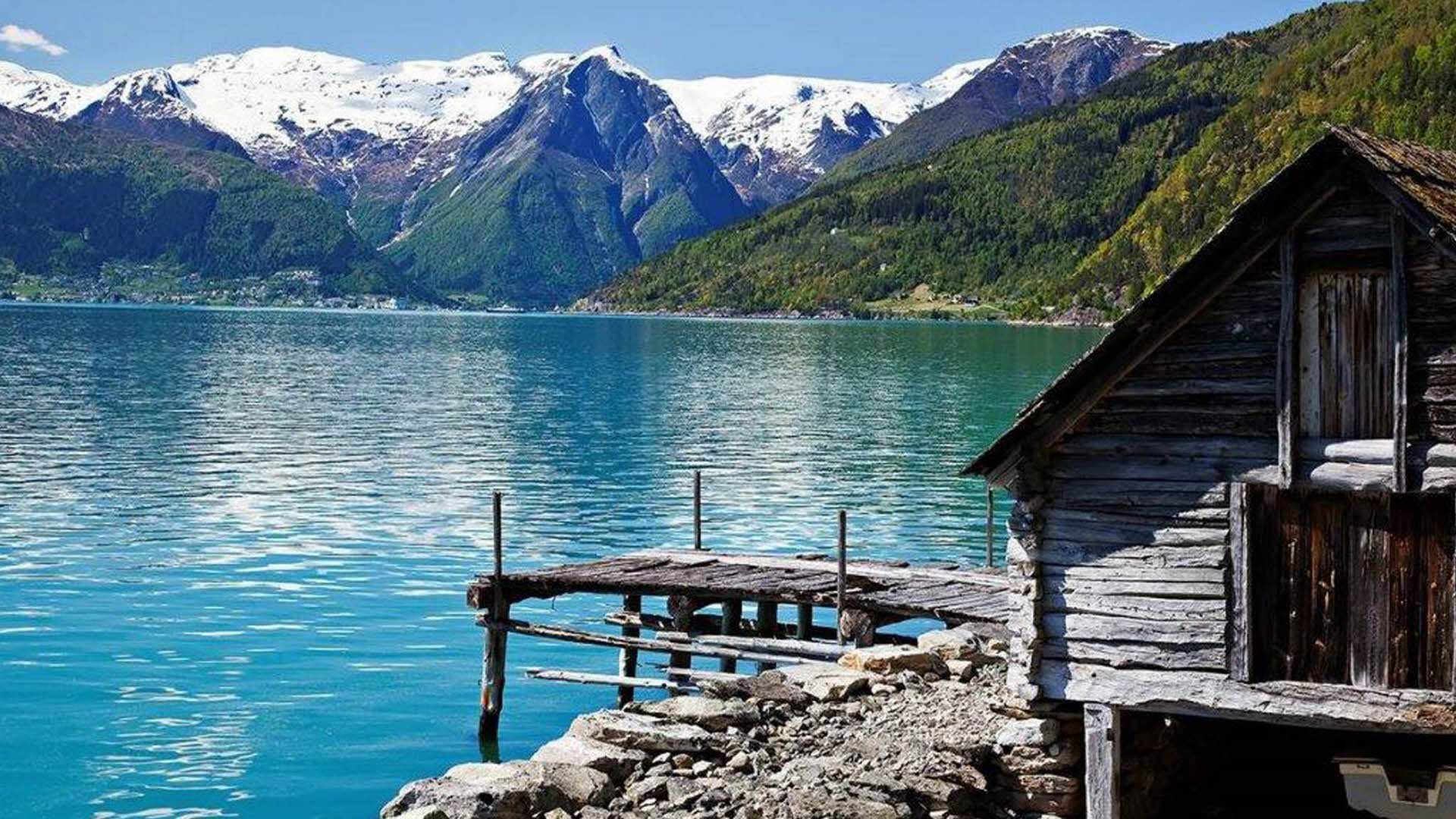 Ice green calm Fjaerlandfjord in summer. Old boathouse and pier in front. Dramatic tall mountains and glacier in distance.
