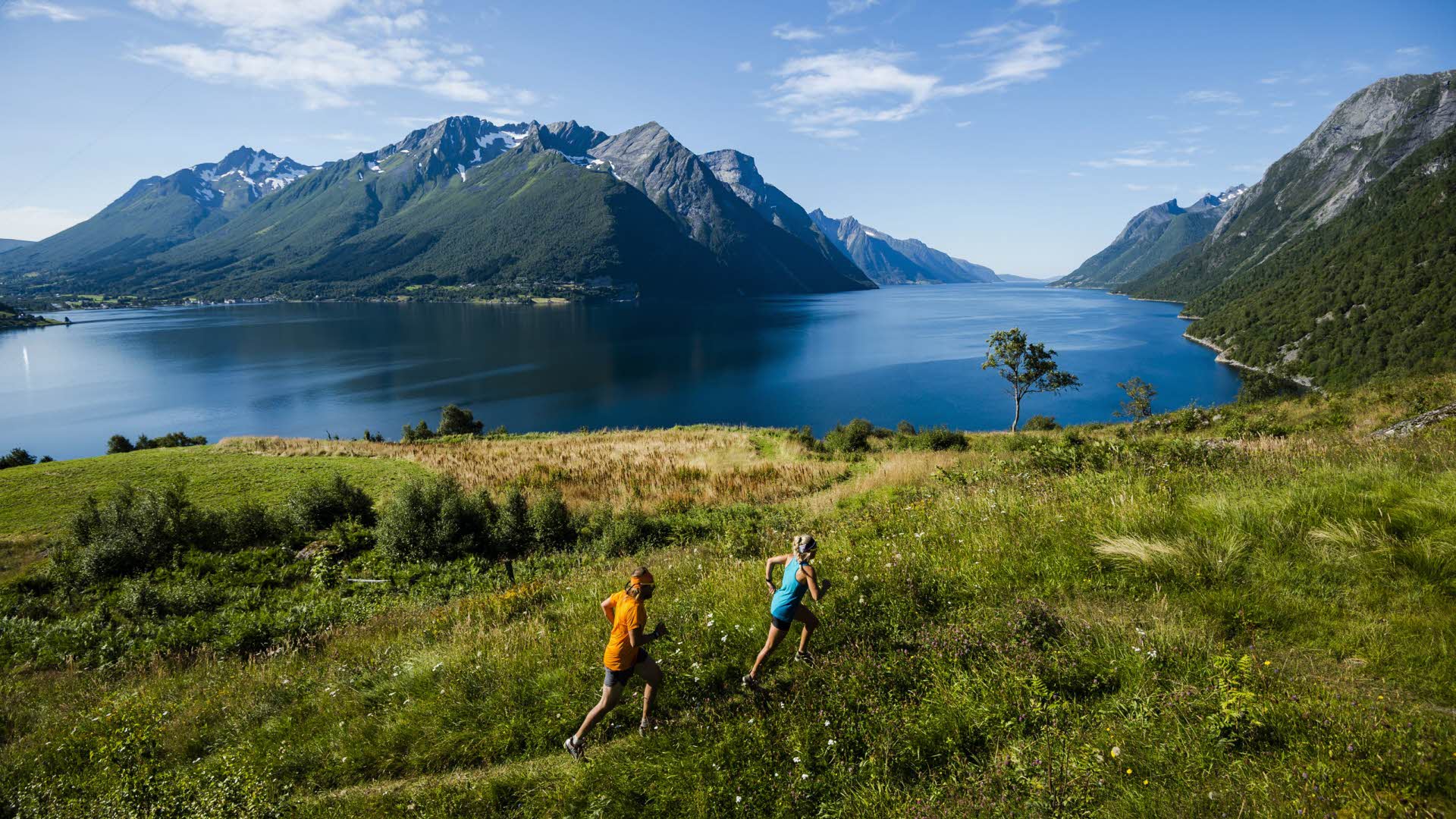 Dos personas caminando en verano por Hjørundfjord con las imponentes montañas de fondo