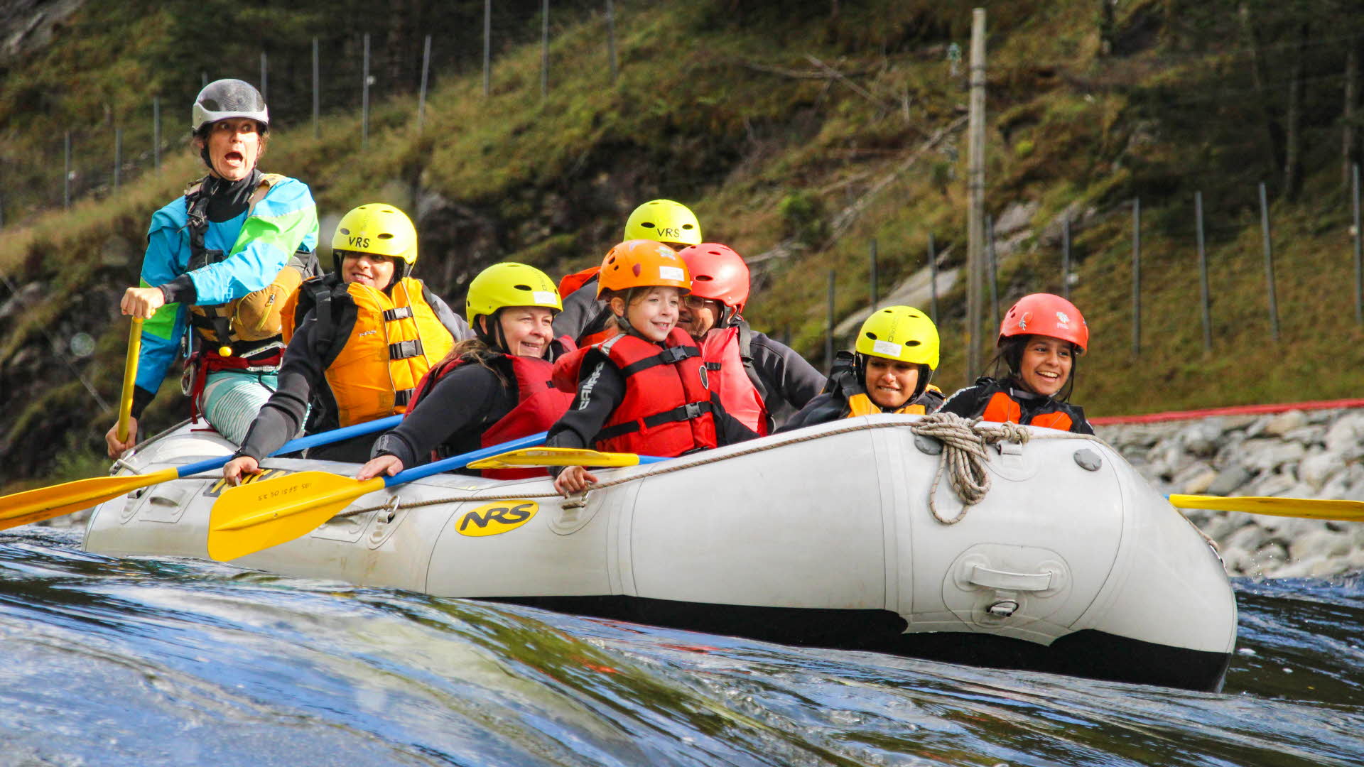 Children in life jackets and helmets sitting in a raft smiling while the guide behind is shouting.