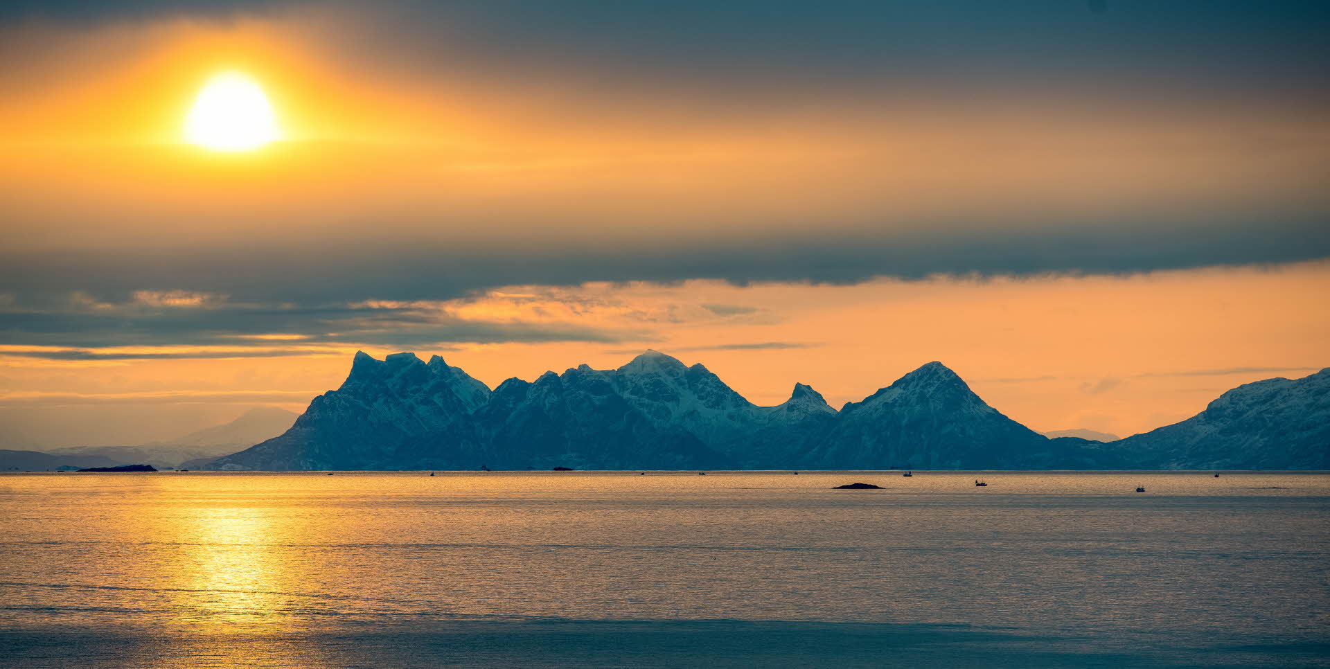 Yellow sun above snow-covered mountains. Calm sea with small boats in front.