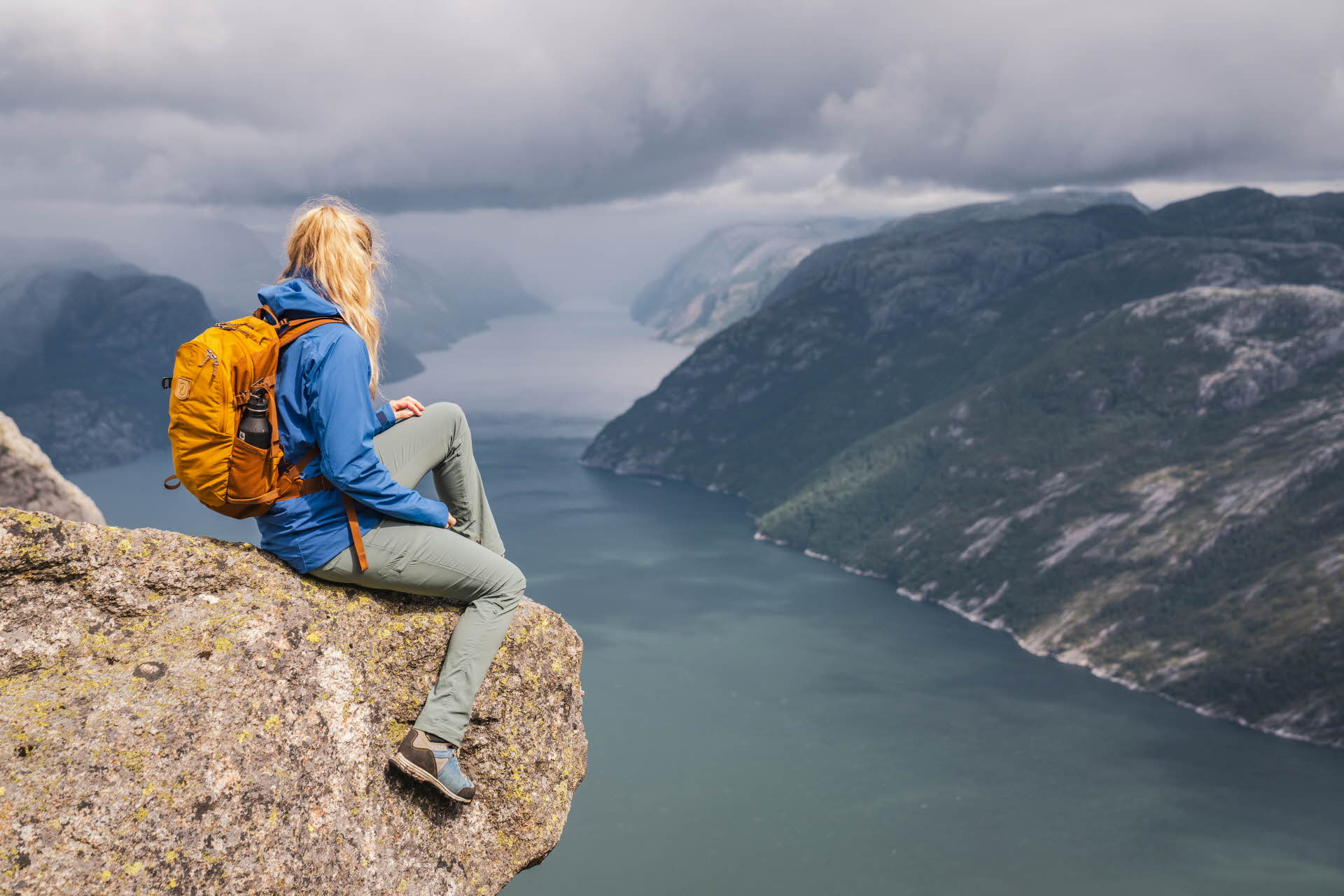 A woman sitting on a rock high above the Lysefjord, looking out at the misty mountains