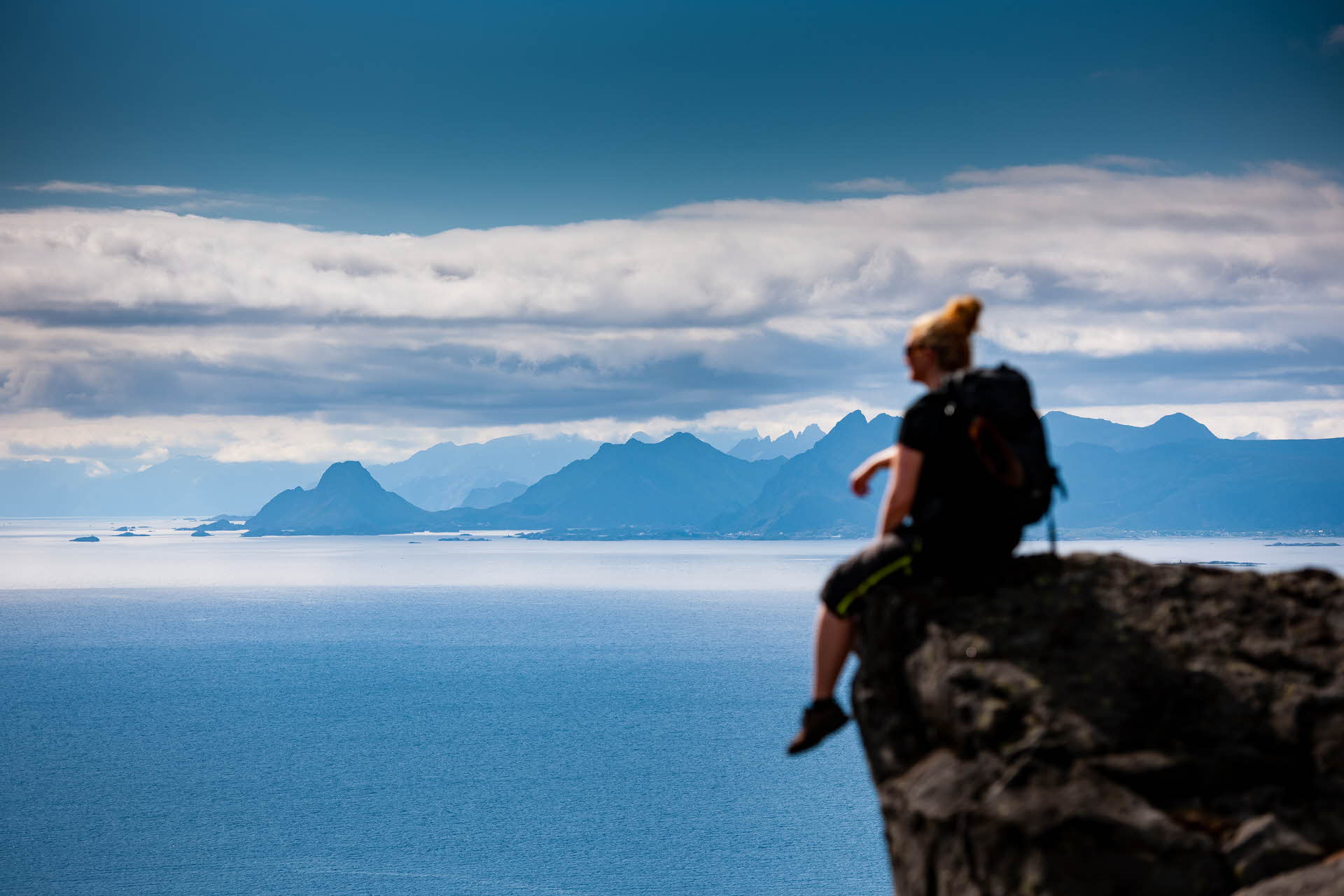 En kvinne sitter på klippe over sjøen. Fjellkjeder i horisonten.