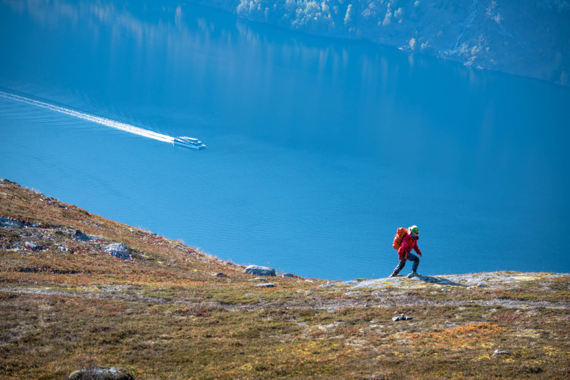 Eine Frau mit roter Jacke und Rucksack wandert oberhalb des Aurlandsfjords bergauf. Auf dem Fjord fährt ein Boot.