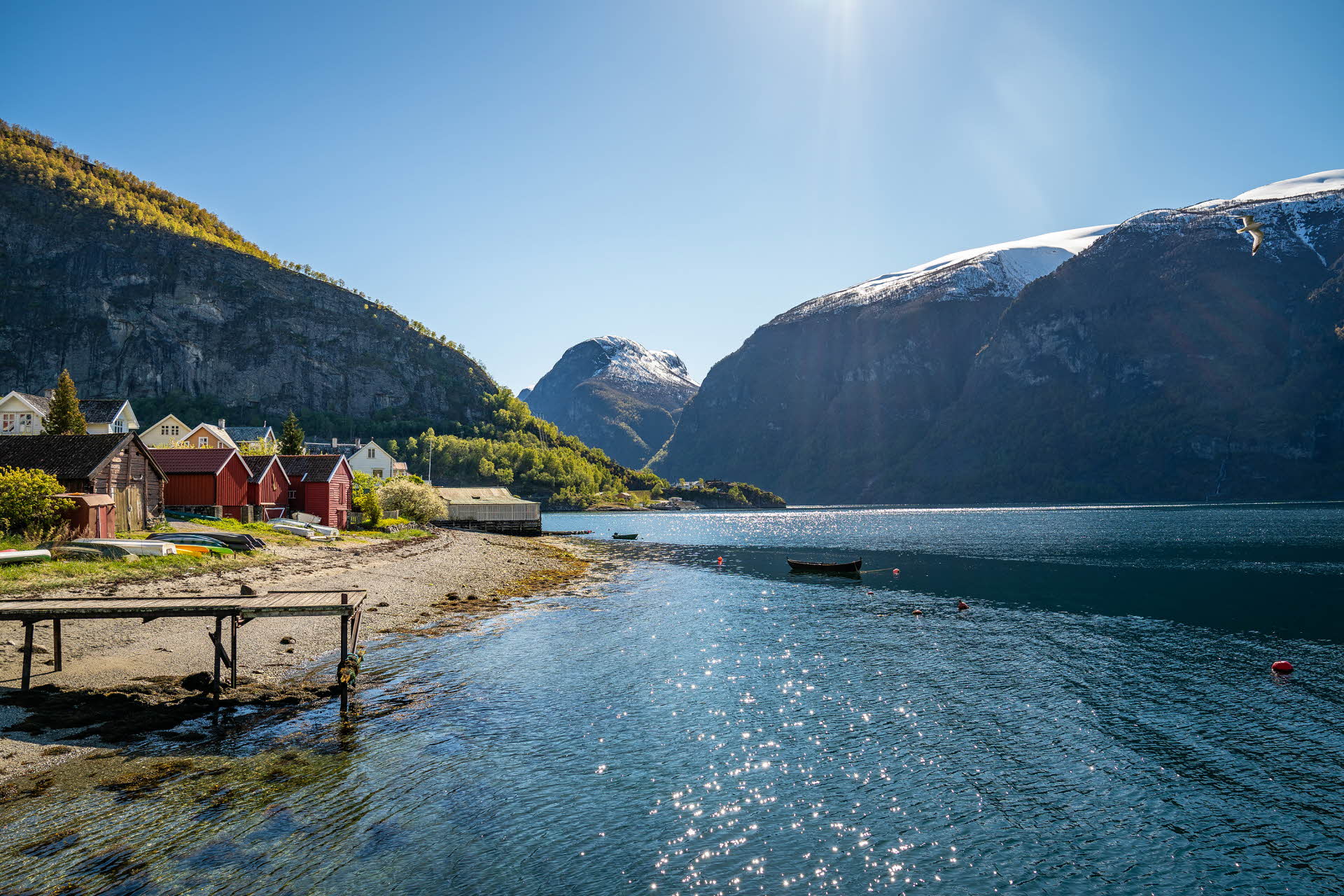 Boat houses at the shoreline by the UNESCO listed Aurlandsfjord in summer. Lush mountain sides with snow-capped mountains