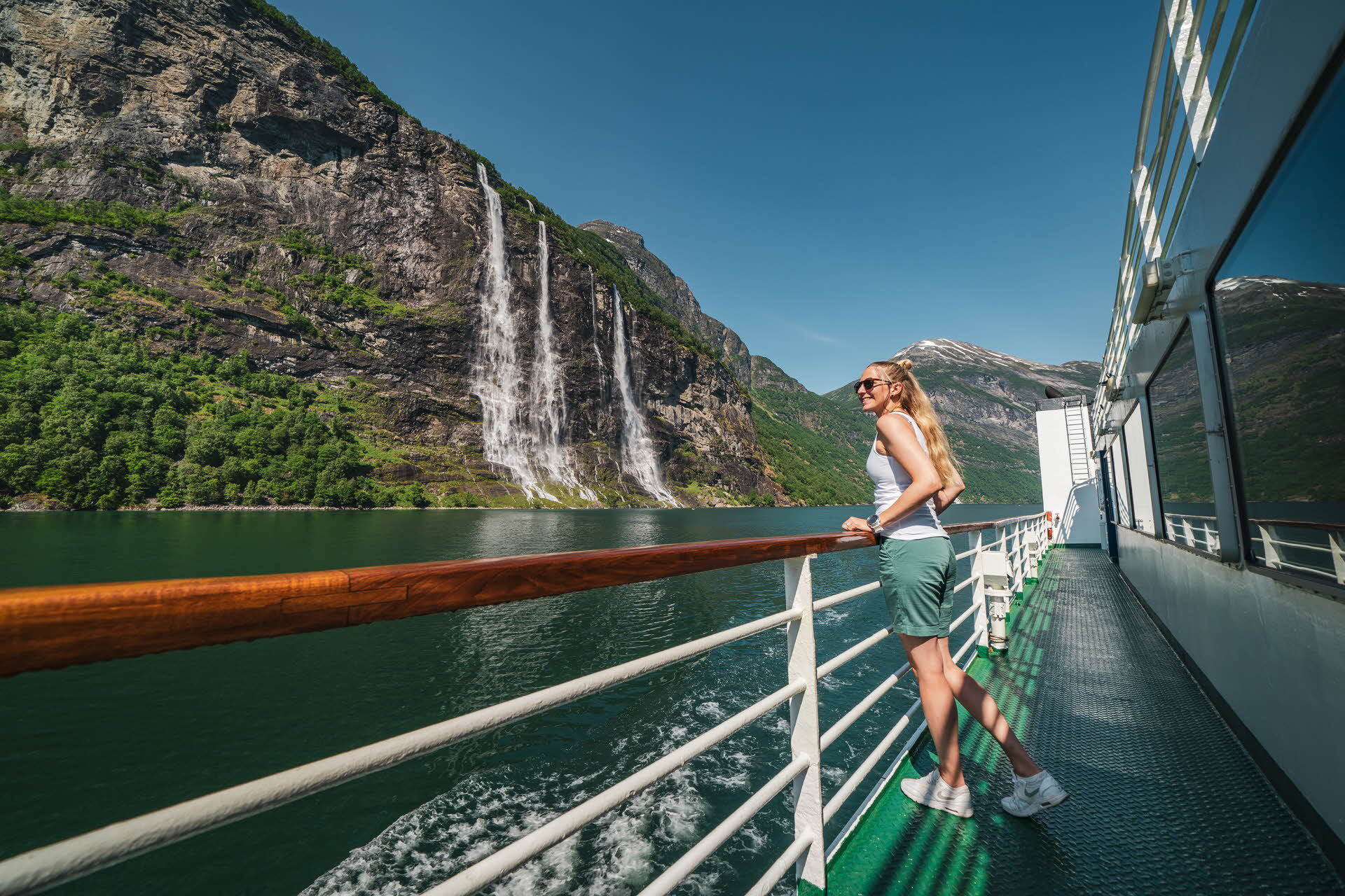 Eine Frau steht an der Reling eines Schiffes auf dem Geirangerfjord, das an einem Wasserfall vorbeigleitet