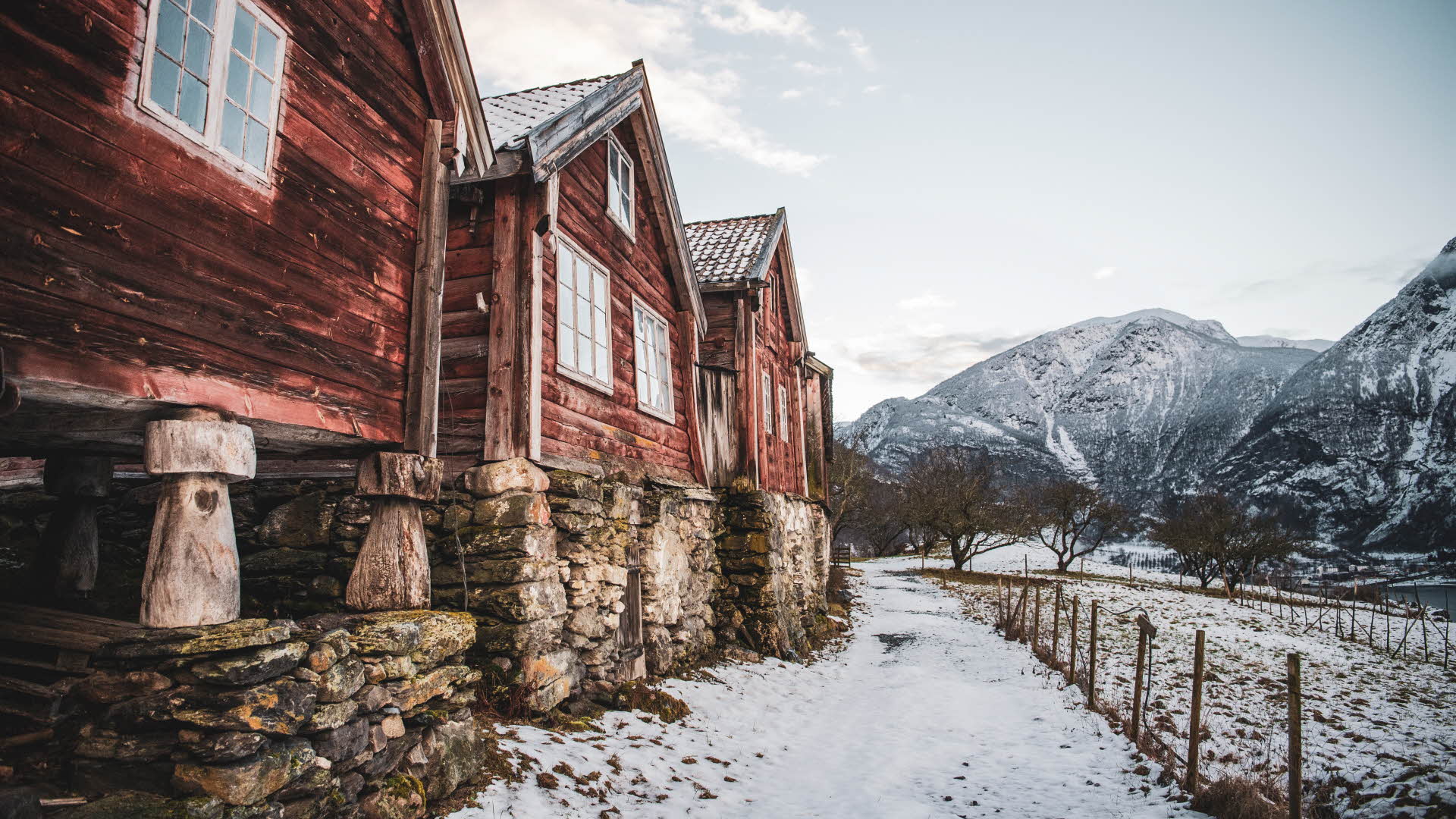 Three old red timber buildings by a field and mountains covered in snow.