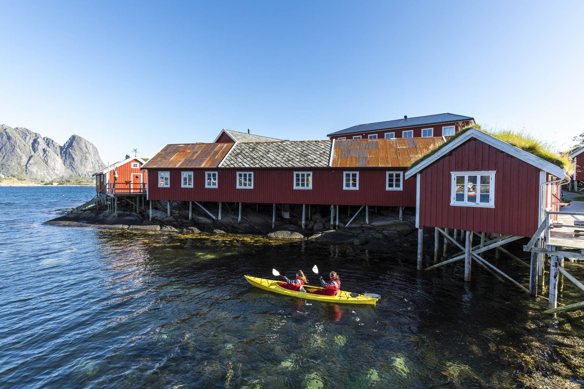 Two people in a tandem kayak on a clear sea next to the red Reine Rorbuer cabins.