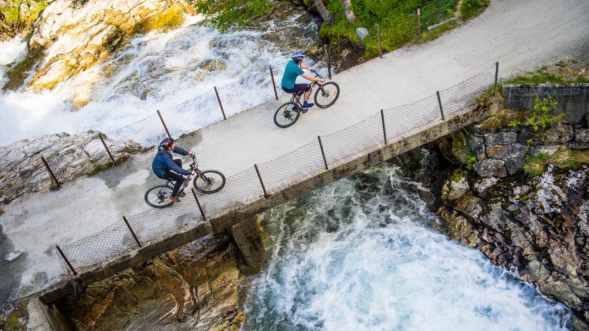 2 cyclists cycling over a bridge in Flåmsdalen, seen from a bird’s eye view