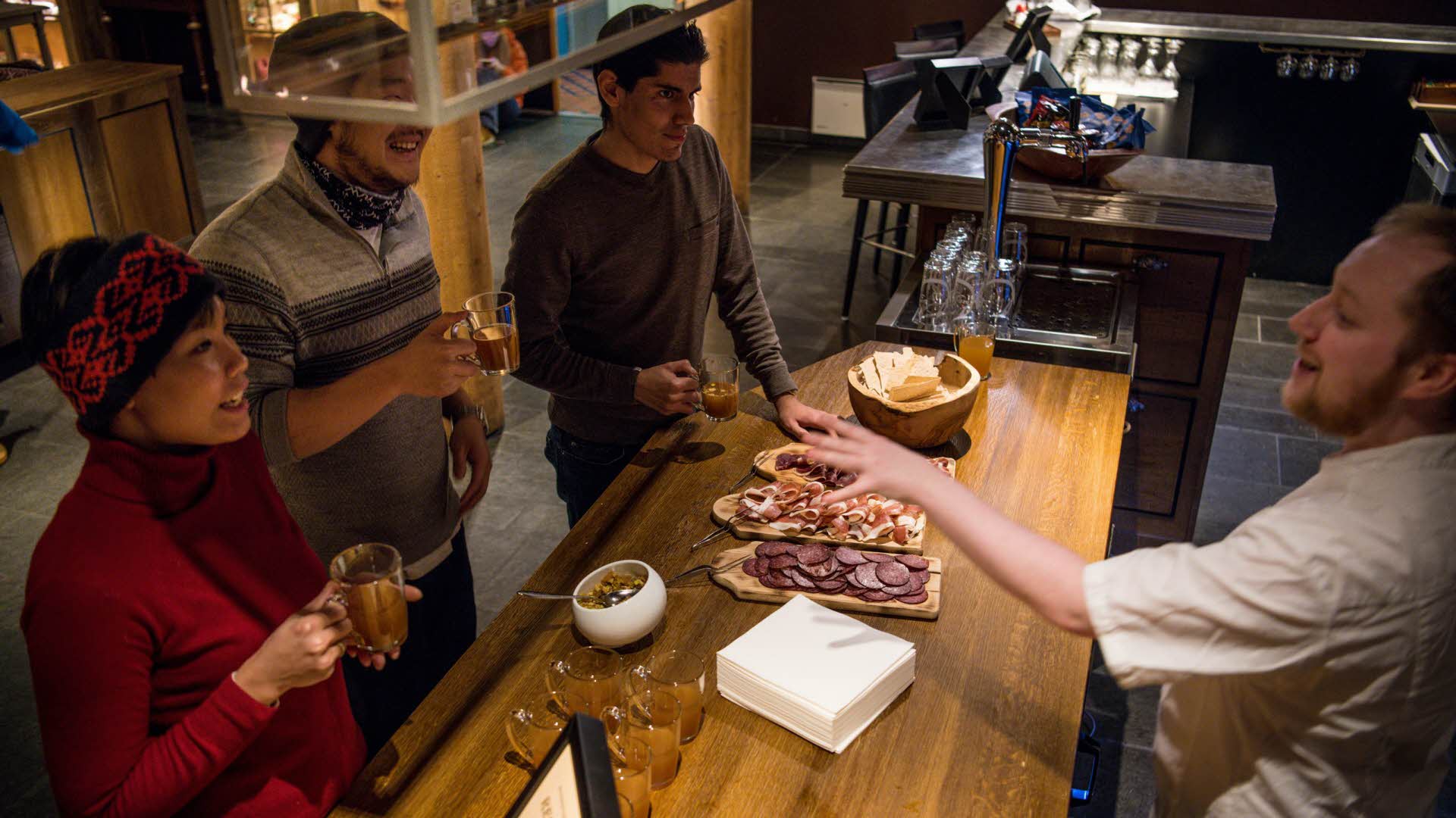 Une femme et deux hommes au bar du hall du Fretheim Hotel, tenant dans leurs mains un verre de vin chaud fait maison et écoutant le chef leur présenter la viande séchée qu’ils sont sur le point de déguster