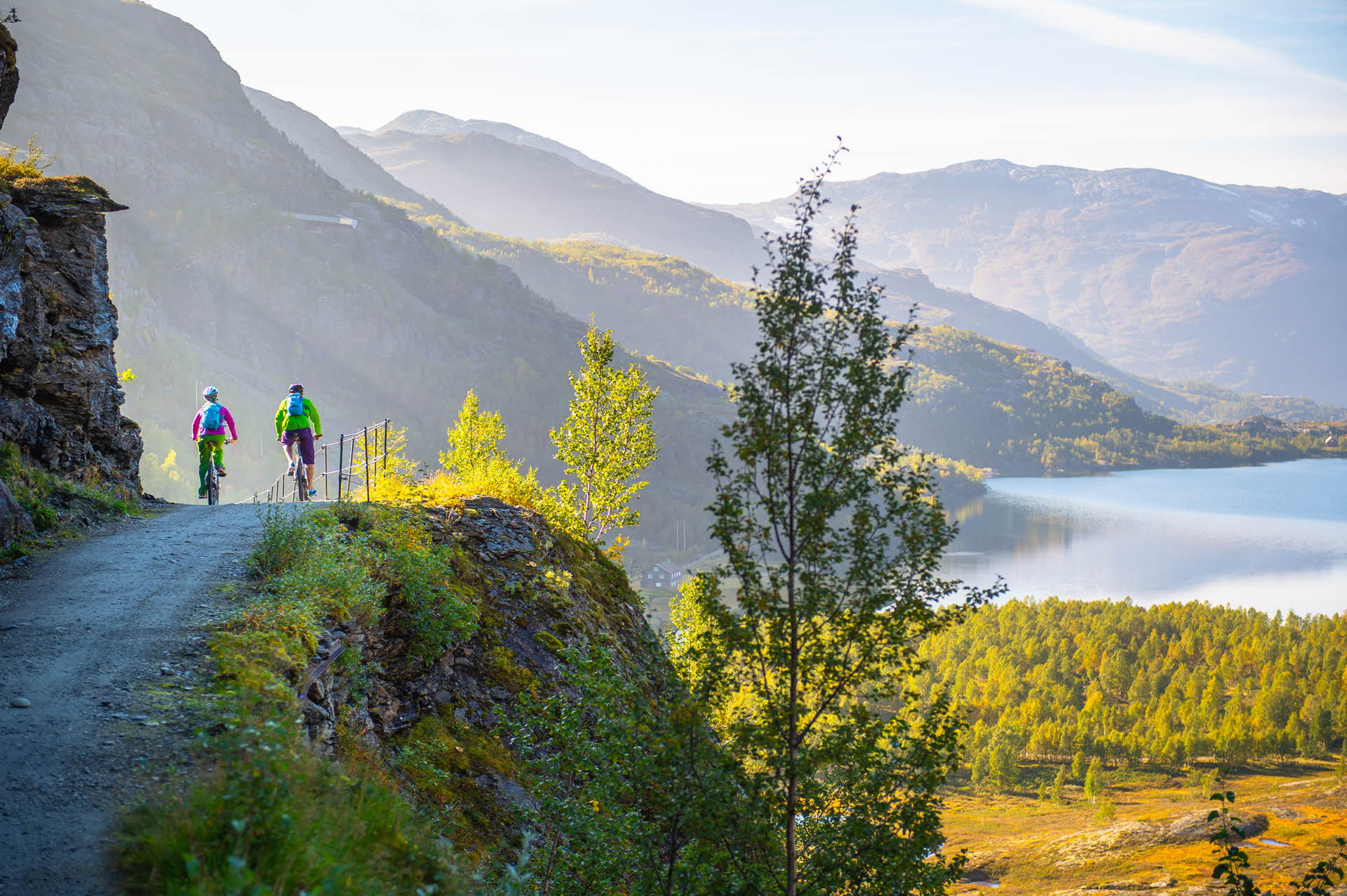 Two cyclists on the Rallarvegen biking beneath mountains towards a lake.