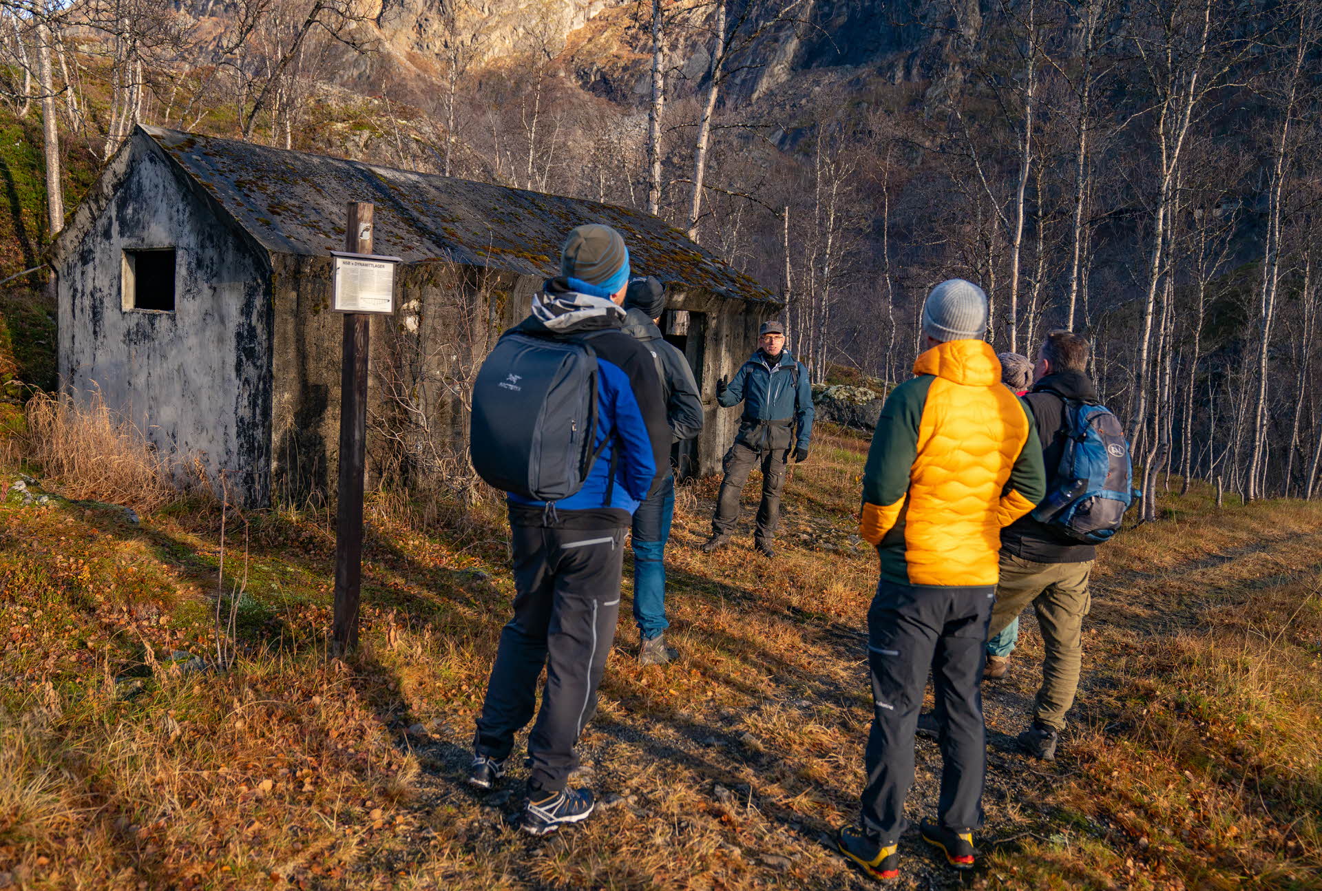 Des hommes debout sur un chemin, près d’une vieille cabane, regardant un autre homme parler. 