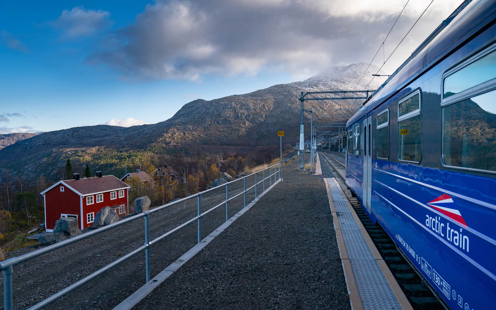 蓝色的北极列车停靠在罗姆巴肯 (Rombak) 车站，近处是红色的建筑和群山