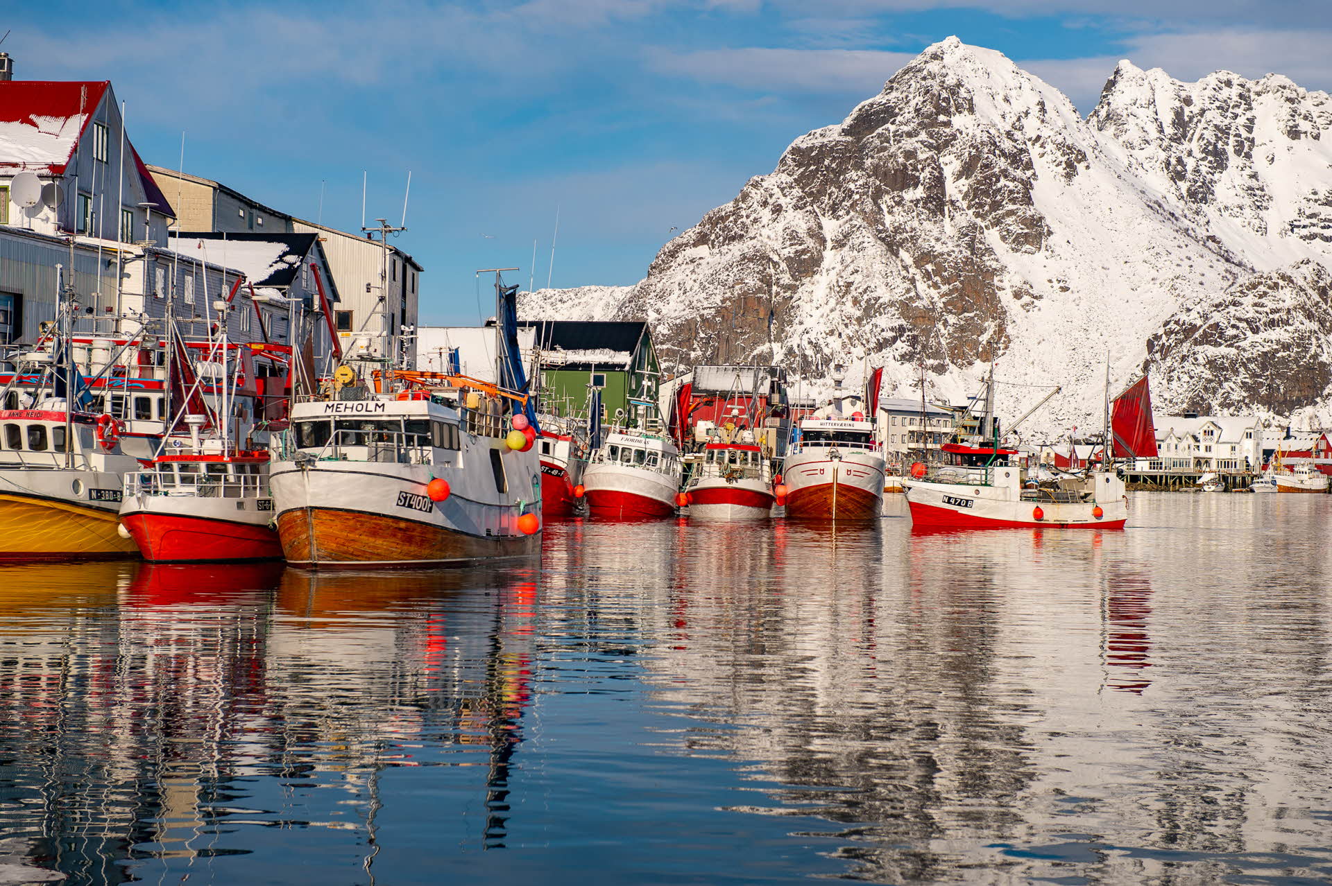 Fishing boats docked at a quay in a village in the Lofoten Islands. Snowy mountains in the background.