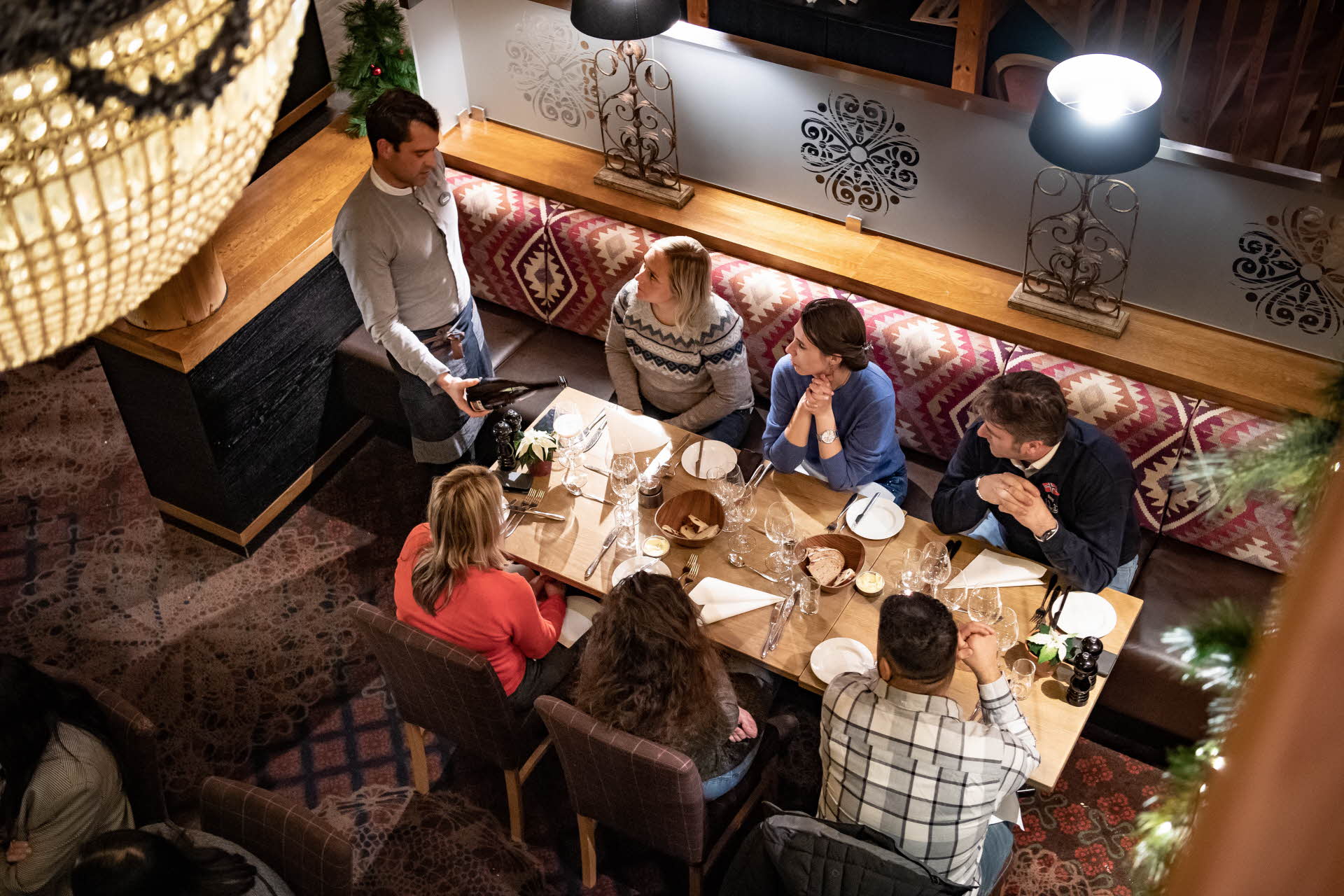 Sechs Gäste an einem Tisch im Restaurant Arven. Ein Kellner serviert ihnen Wein. Von oben gesehen, mit einem Kronleuchter im Vordergrund.