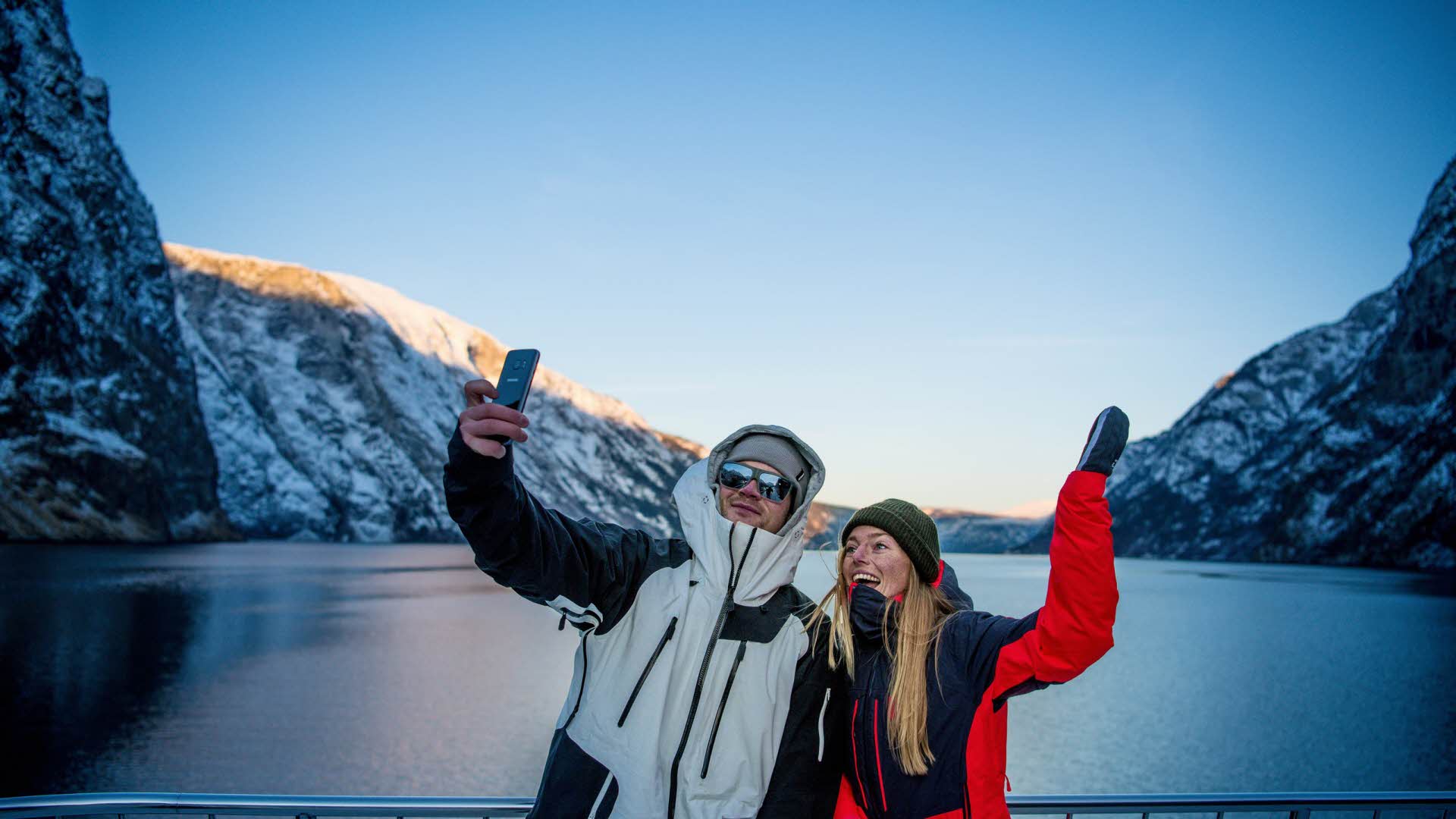 En gutt og en jente tar selfie fra en båt på Nærøyfjorden i blått vinterlys. 