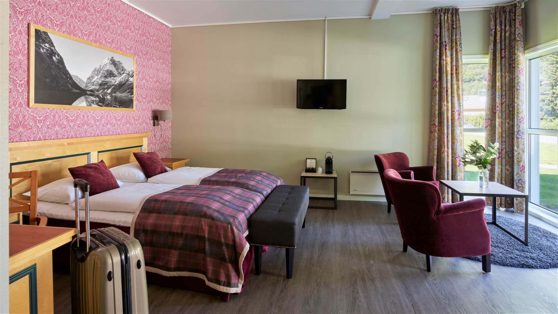 Junior suite at Fretheim Hotel showing twin beds, espresso machine and two red lounge chairs facing the windows by the garden. Suitcase standing in front. 