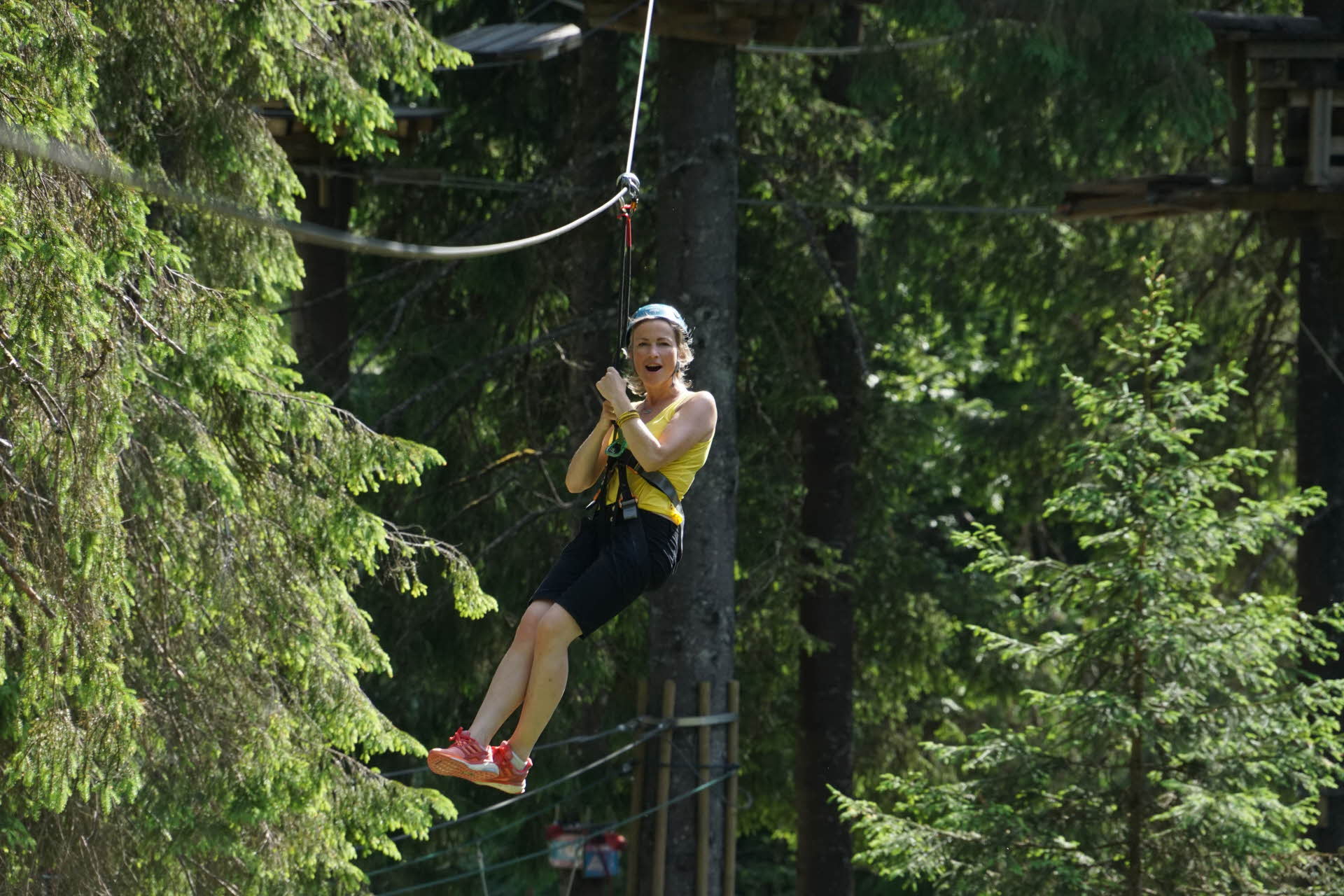 A woman in summer clothes hanging from a zipline and screaming. Surrounded by a green forest.