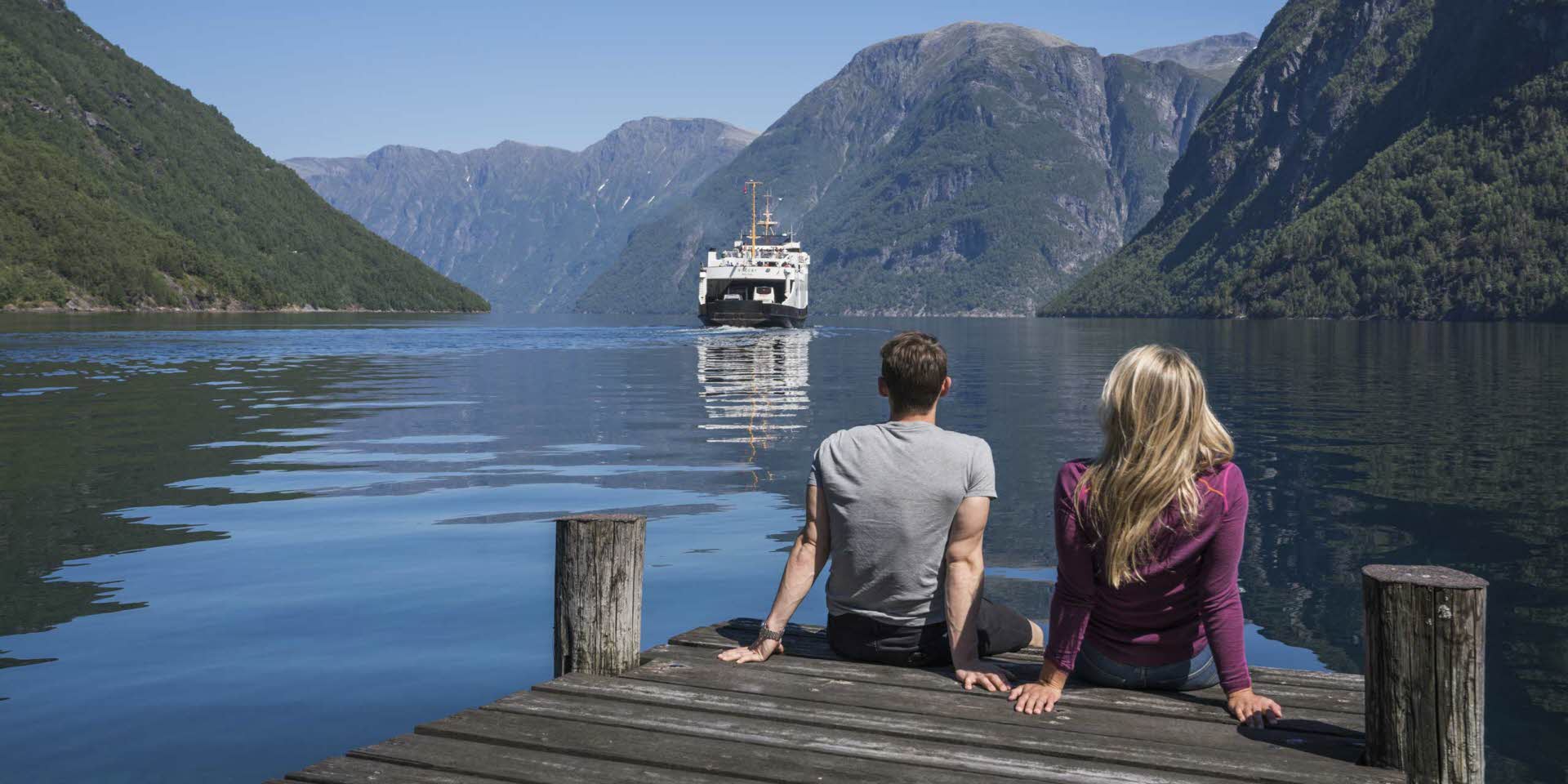 Ein Pärchen sitzt auf einem Holzsteg mit Blick auf den Geirangerfjord, während die Fähre näher kommt