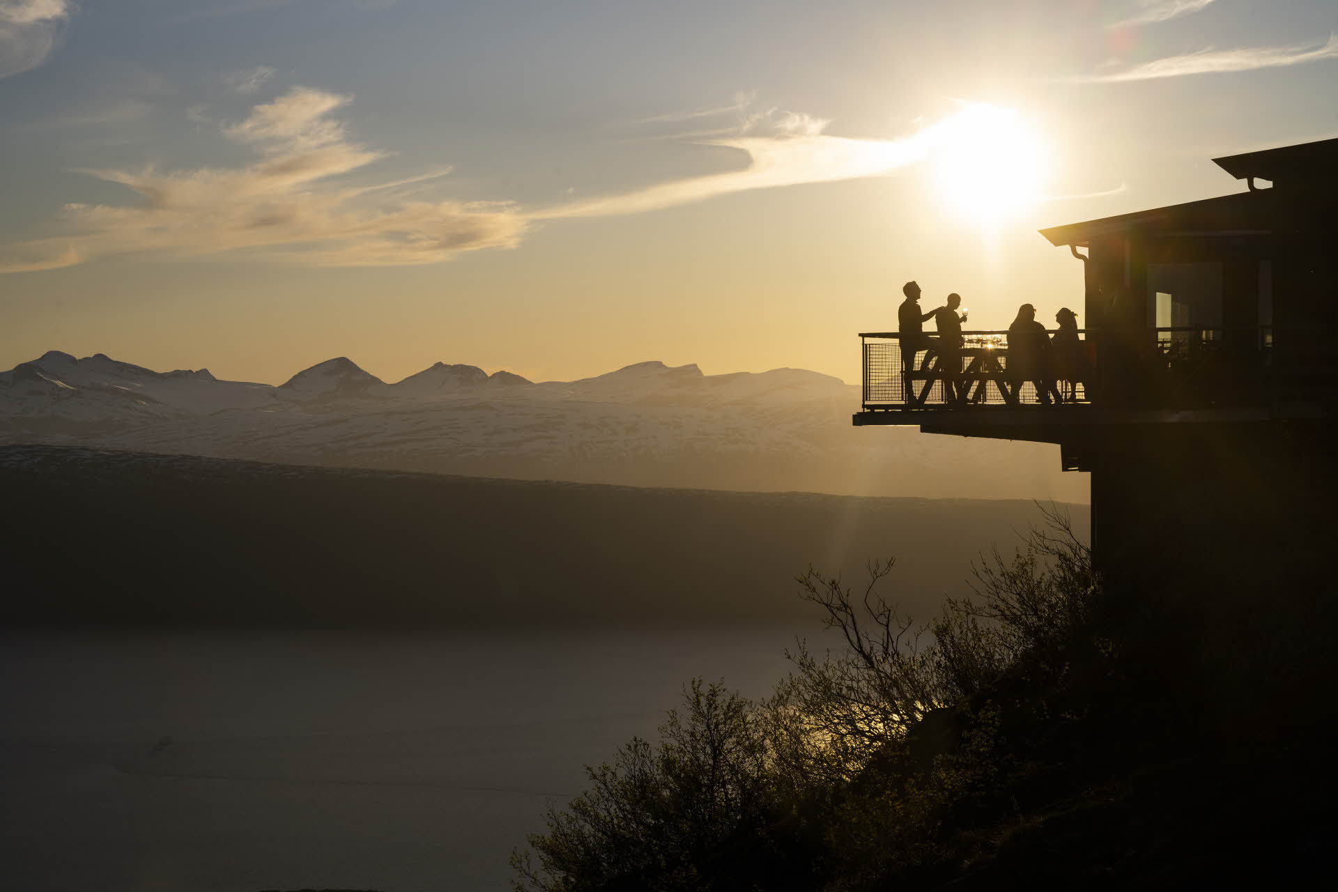 Des personnes se tiennent debout sur le balcon du restaurant Narvikfjellet au soleil de minuit. Les fjords et les montagnes sont visibles à l’arrière-plan.