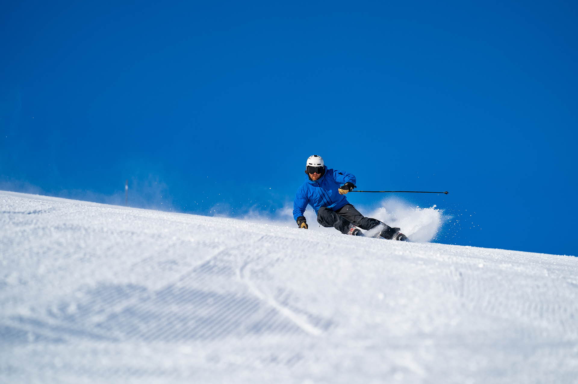 一个穿蓝色夹克滑雪的人在米克达林完美的滑雪坡道上铲起雪花。后面的天很蓝。 