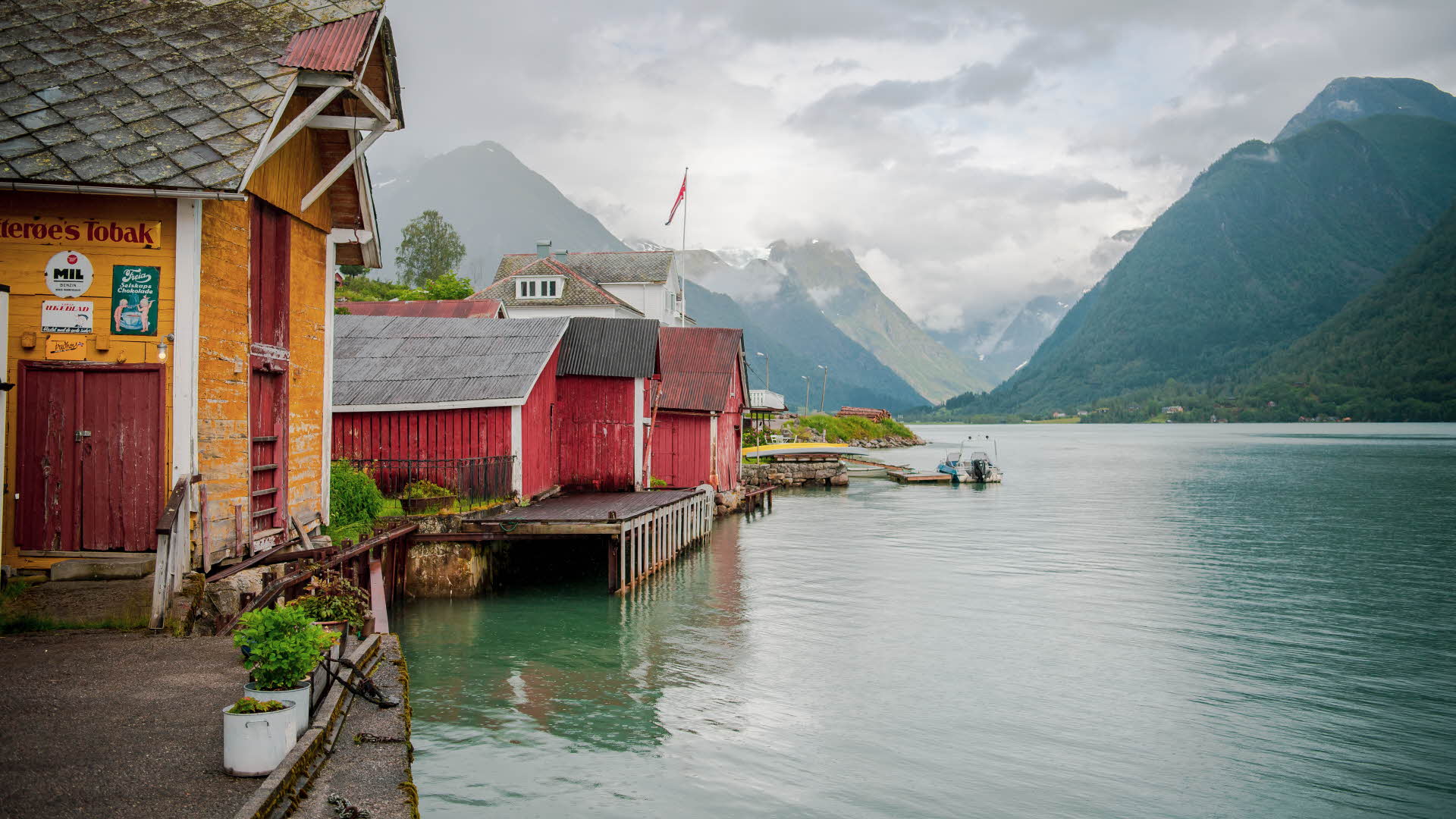 One yellow and two red boathouses along the Fjærlandsfjord, with grey clouds over the mountains