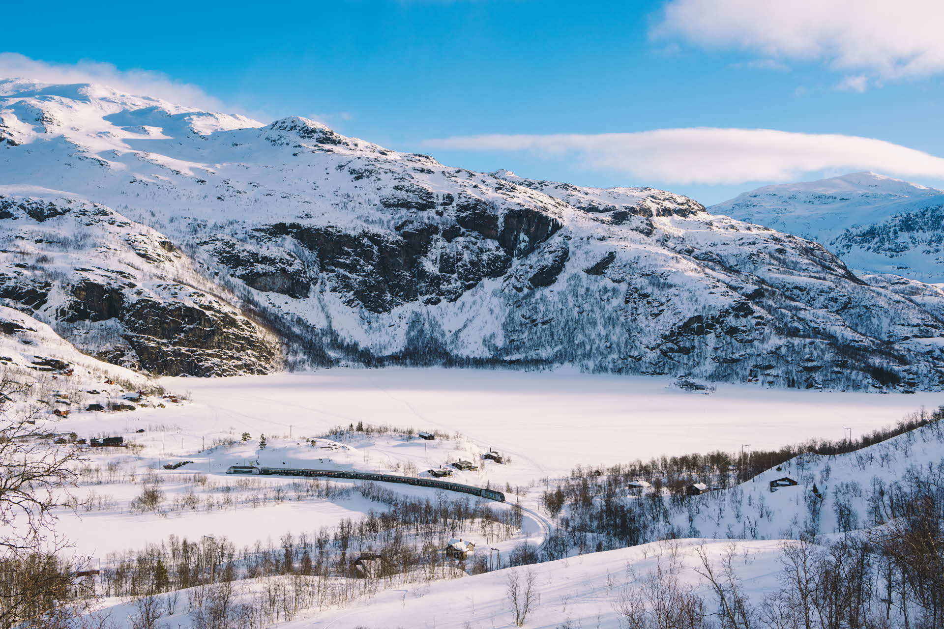 Flåmsbana running through a winter landscape with a frozen lake and snow-covered mountains. There is sunlight on the peaks beneath a clear blue sky. 