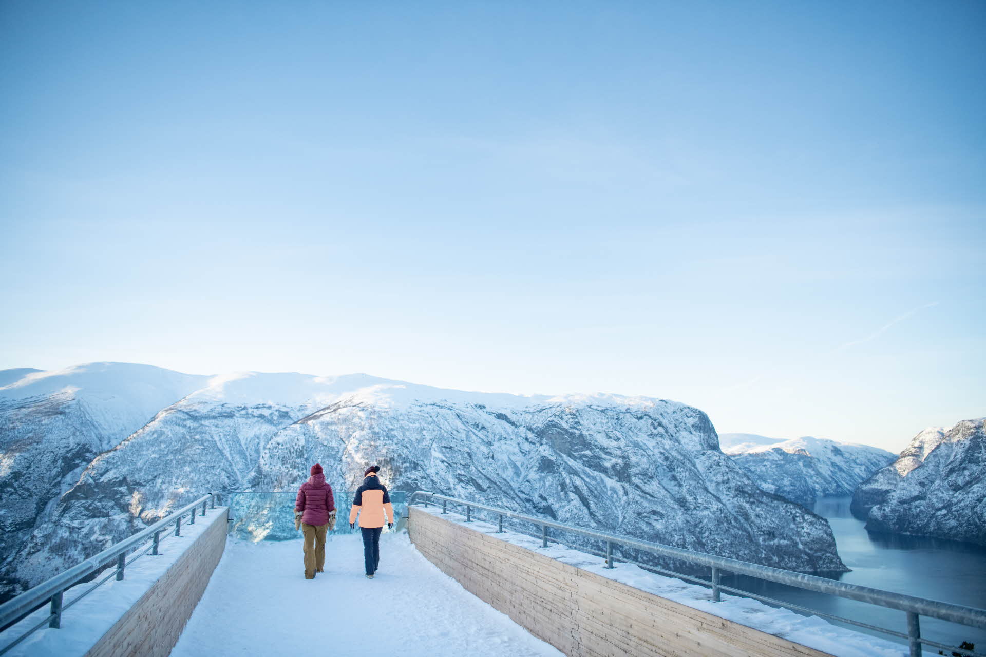 Two people in winter outfit walking on snow on Stegastein Viewpoint surrounded by winter landscape. Photo. 