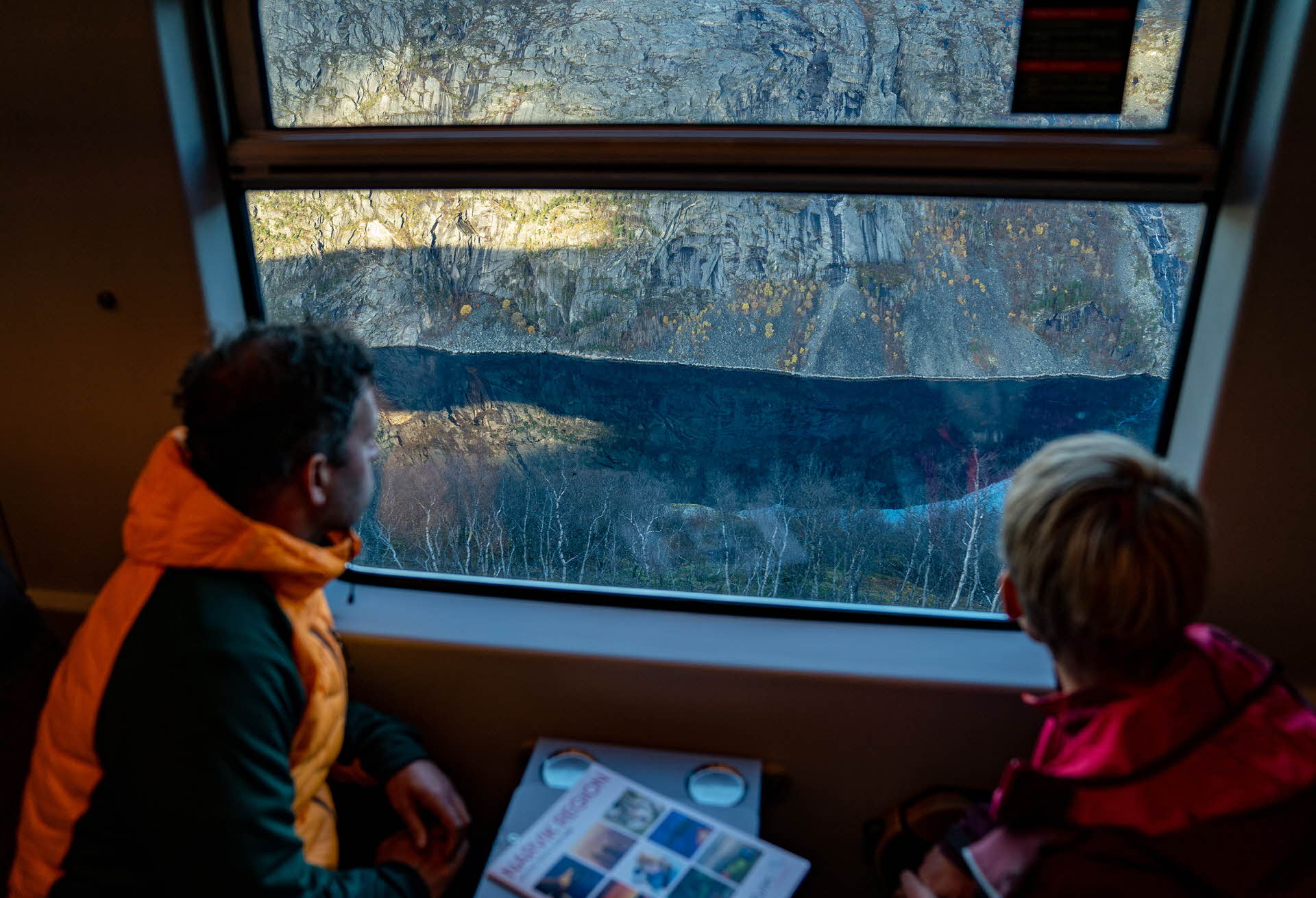 Un hombre y una mujer sentados uno frente al otro en un tren mirando por la ventana hacia Rombaksbotn